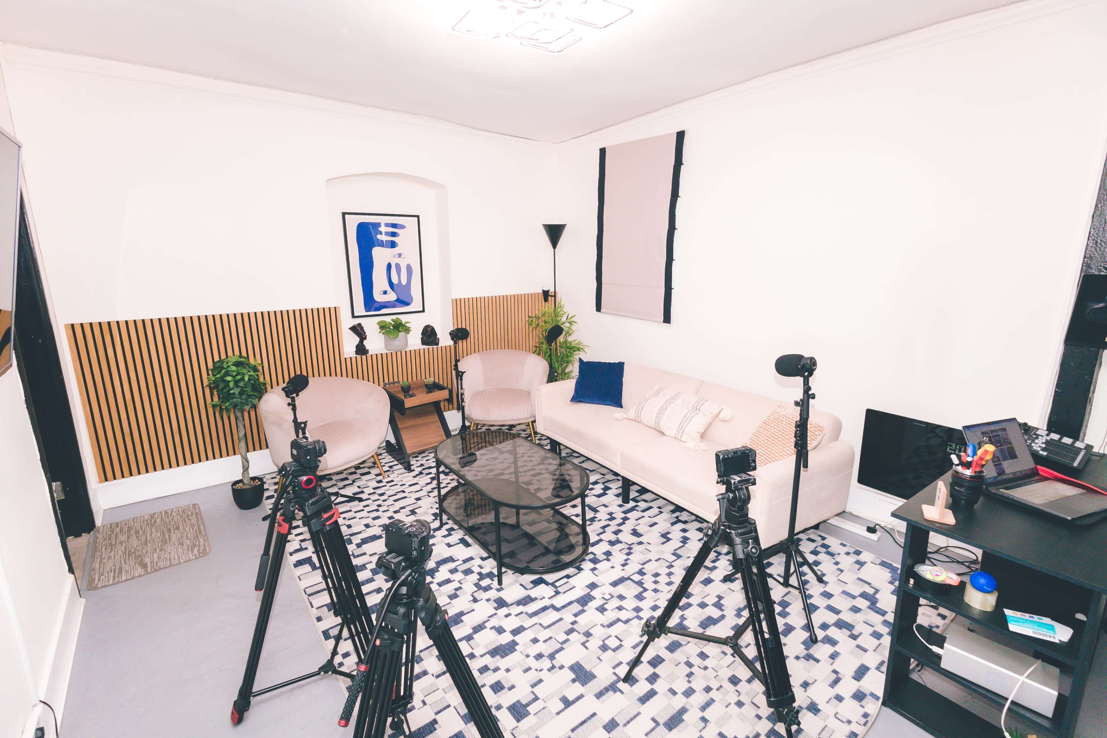 A brightly lit room features two armchairs and a sofa around a glass coffee table, with several cameras positioned on tripods and plants placed throughout.