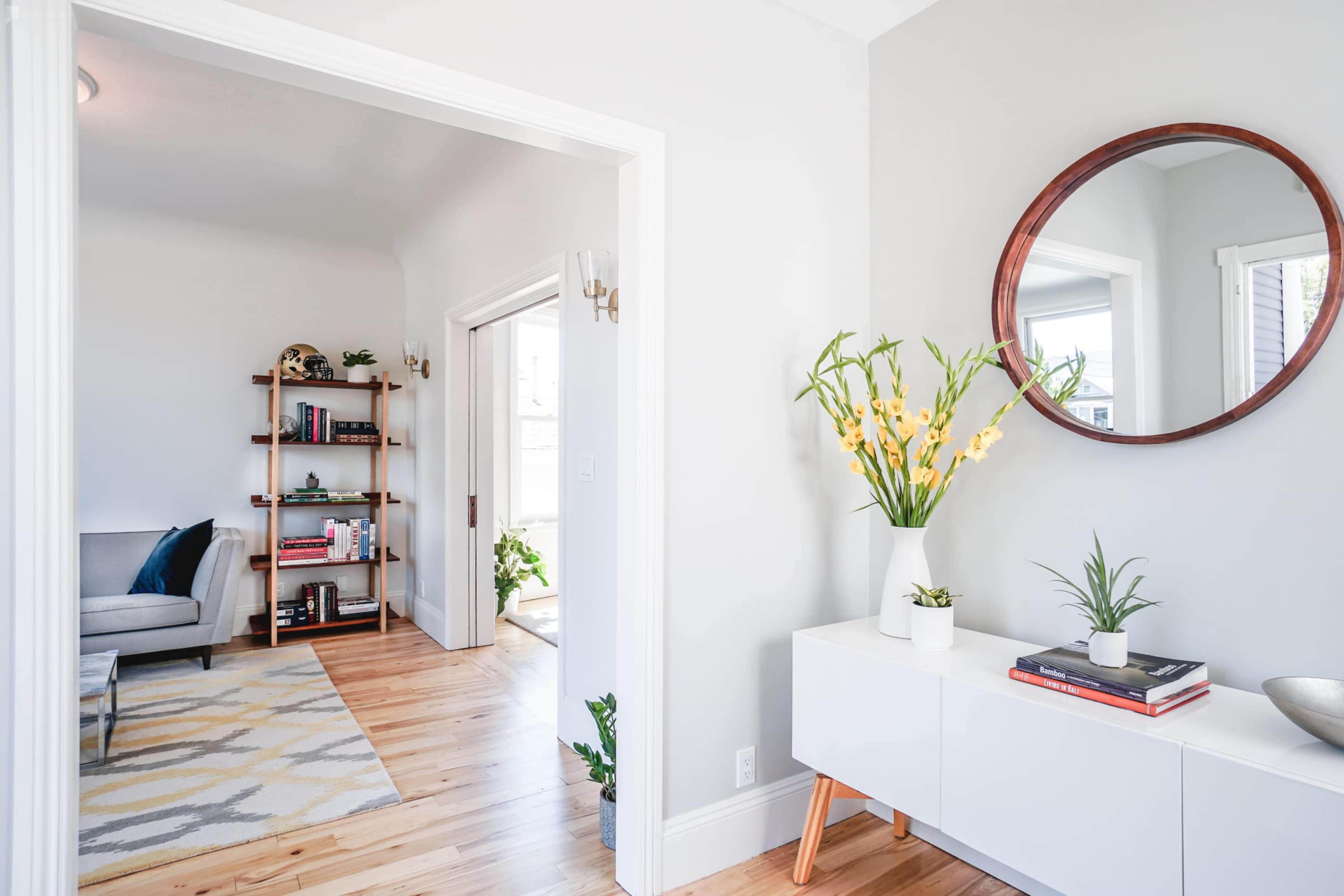 A well-lit entryway that leads to a living area, featuring a round mirror above a white sideboard adorned with plants and books.