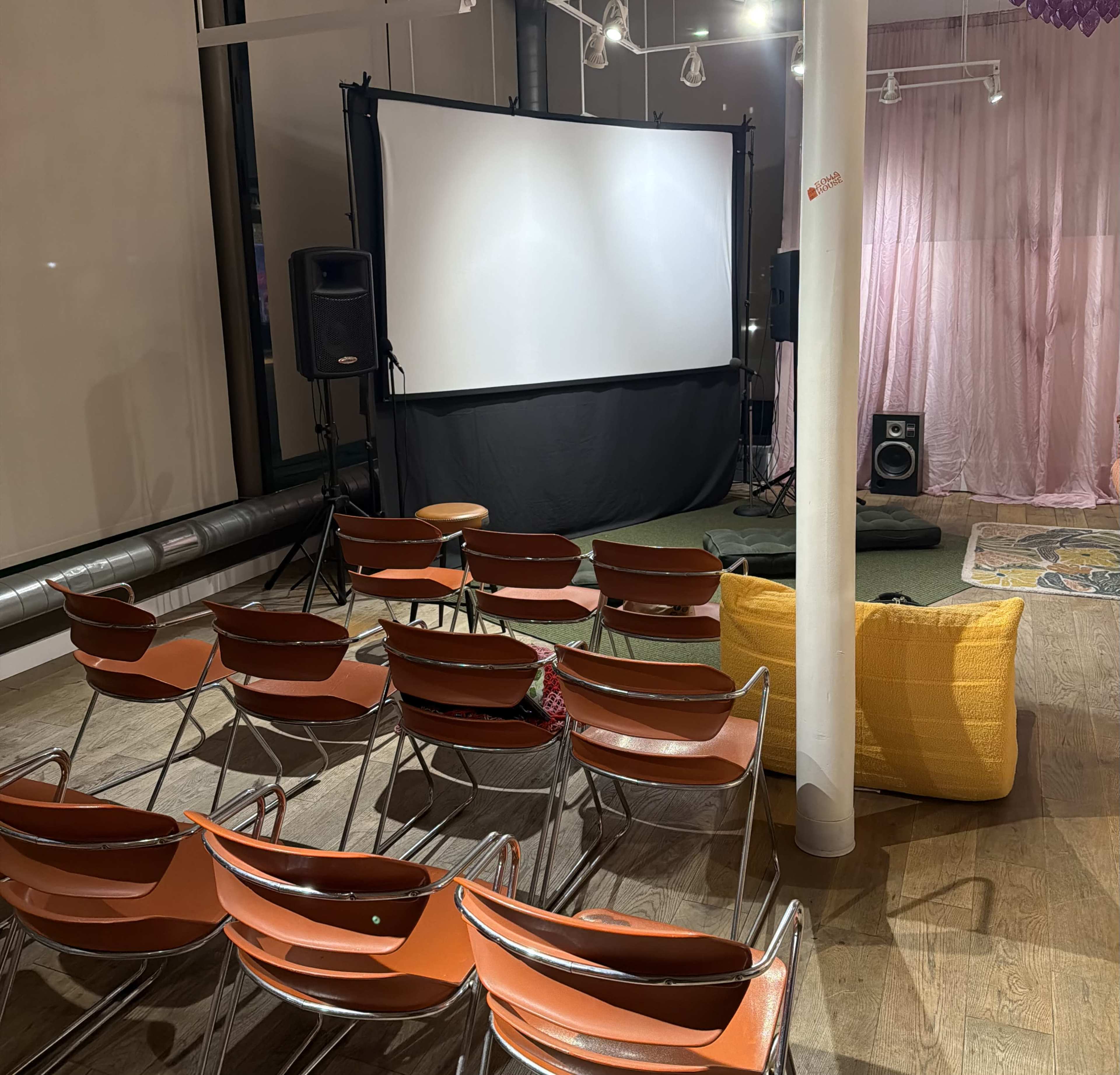 The image shows a small indoor space set up for a presentation or screening, featuring a projector screen, rows of orange chairs, and decorative elements like a pink curtain and speakers.