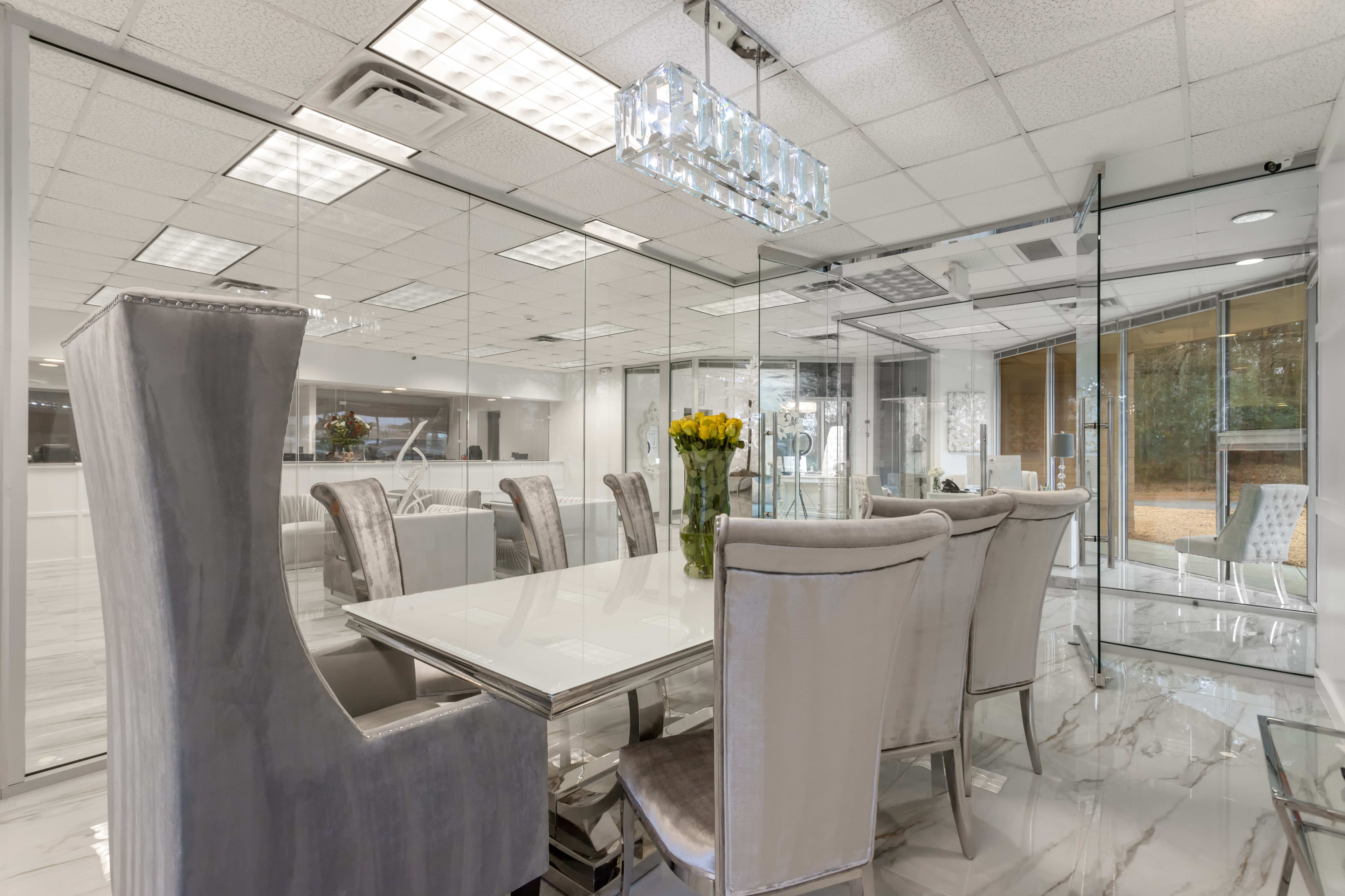 A modern conference room with a glass-enclosed design, featuring a white table and plush, light gray chairs arranged around it, with a floral centerpiece and ample lighting from the ceiling.