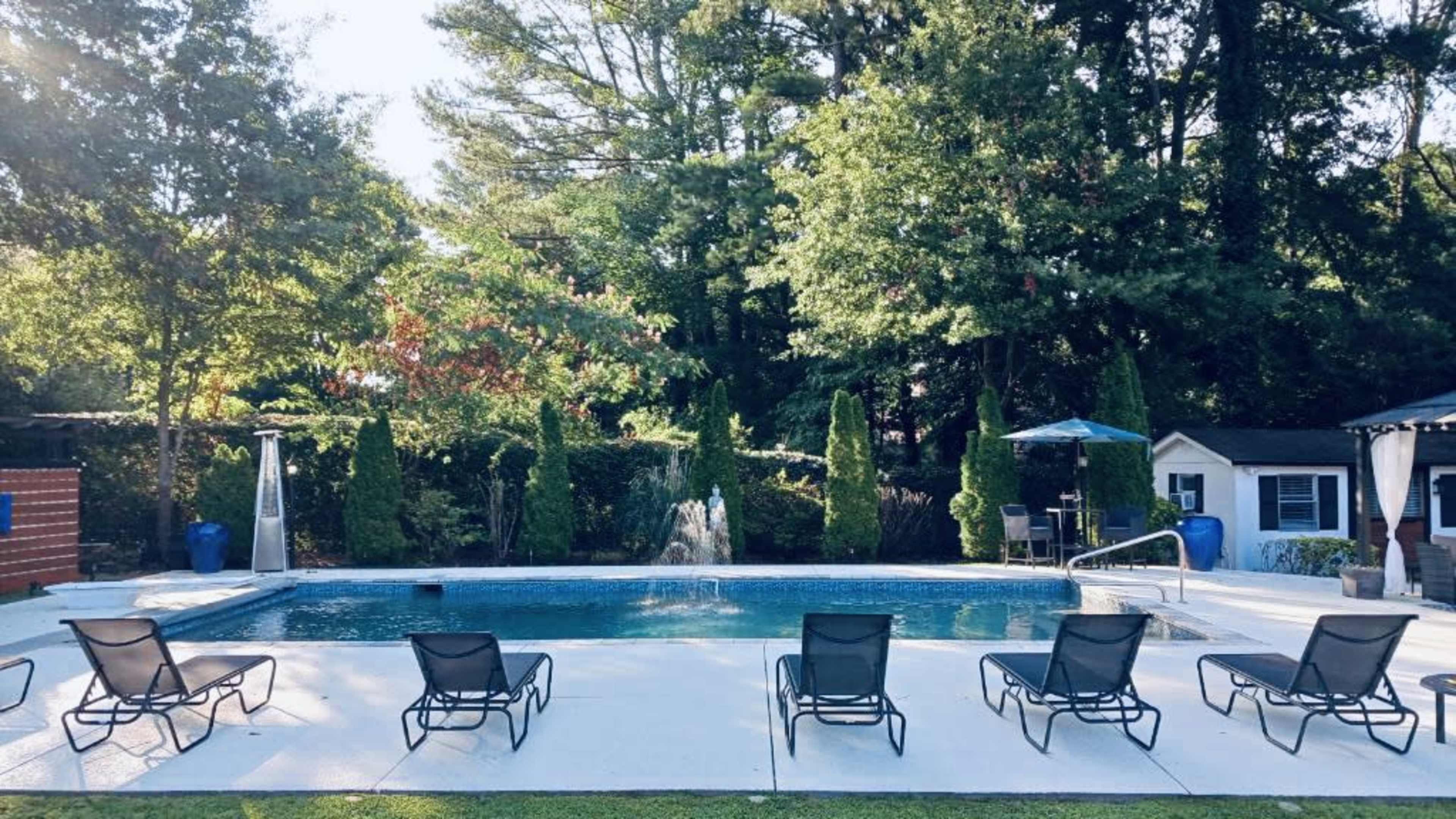 A swimming pool surrounded by lounge chairs, with trees and a small fountain in the background.