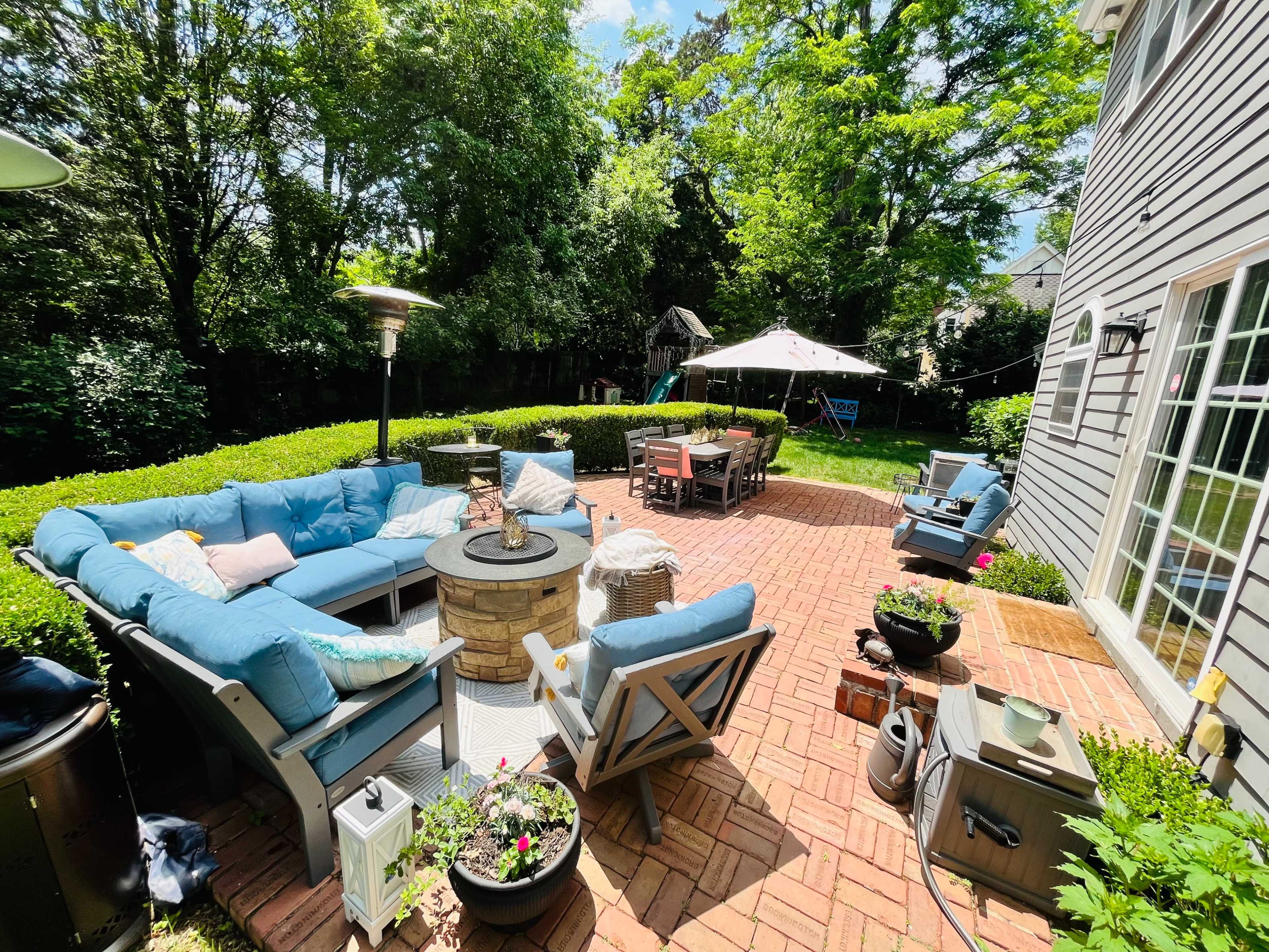 A backyard patio features a blue sectional sofa arranged around a stone fire pit, surrounded by greenery and outdoor dining furniture under a canopy.
