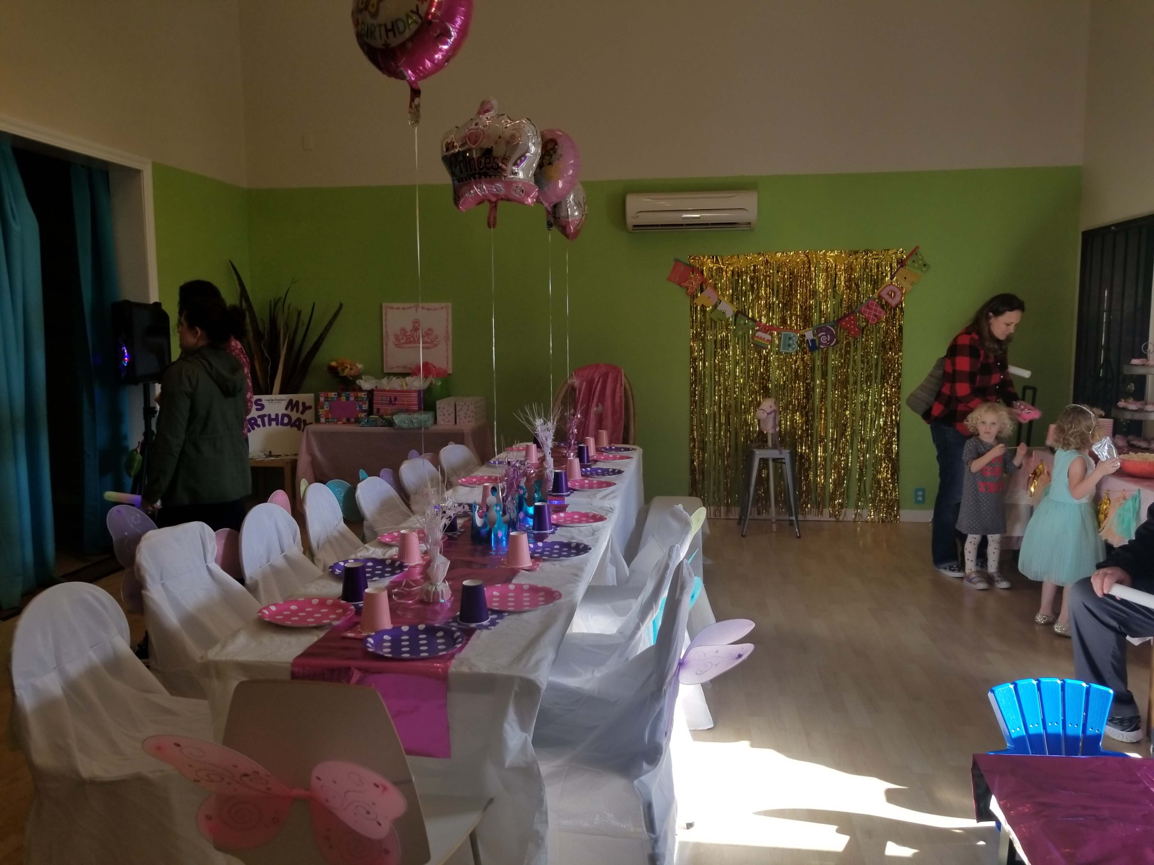 A decorated indoor space set up for a birthday party, featuring a long table with colorful decorations, balloons, and several guests.