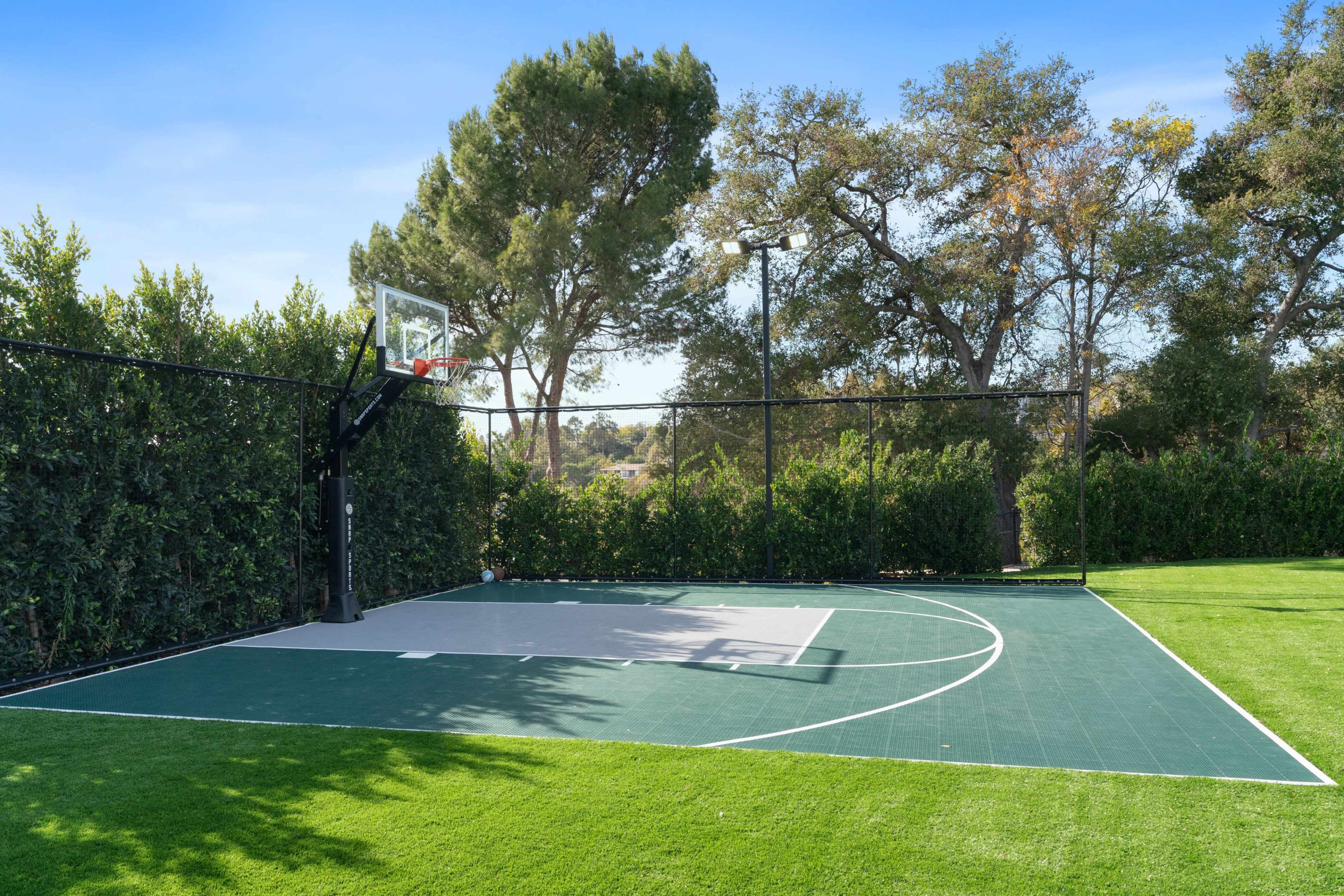 The image shows a basketball court with a hoop surrounded by tall trees and a well-maintained grassy area.