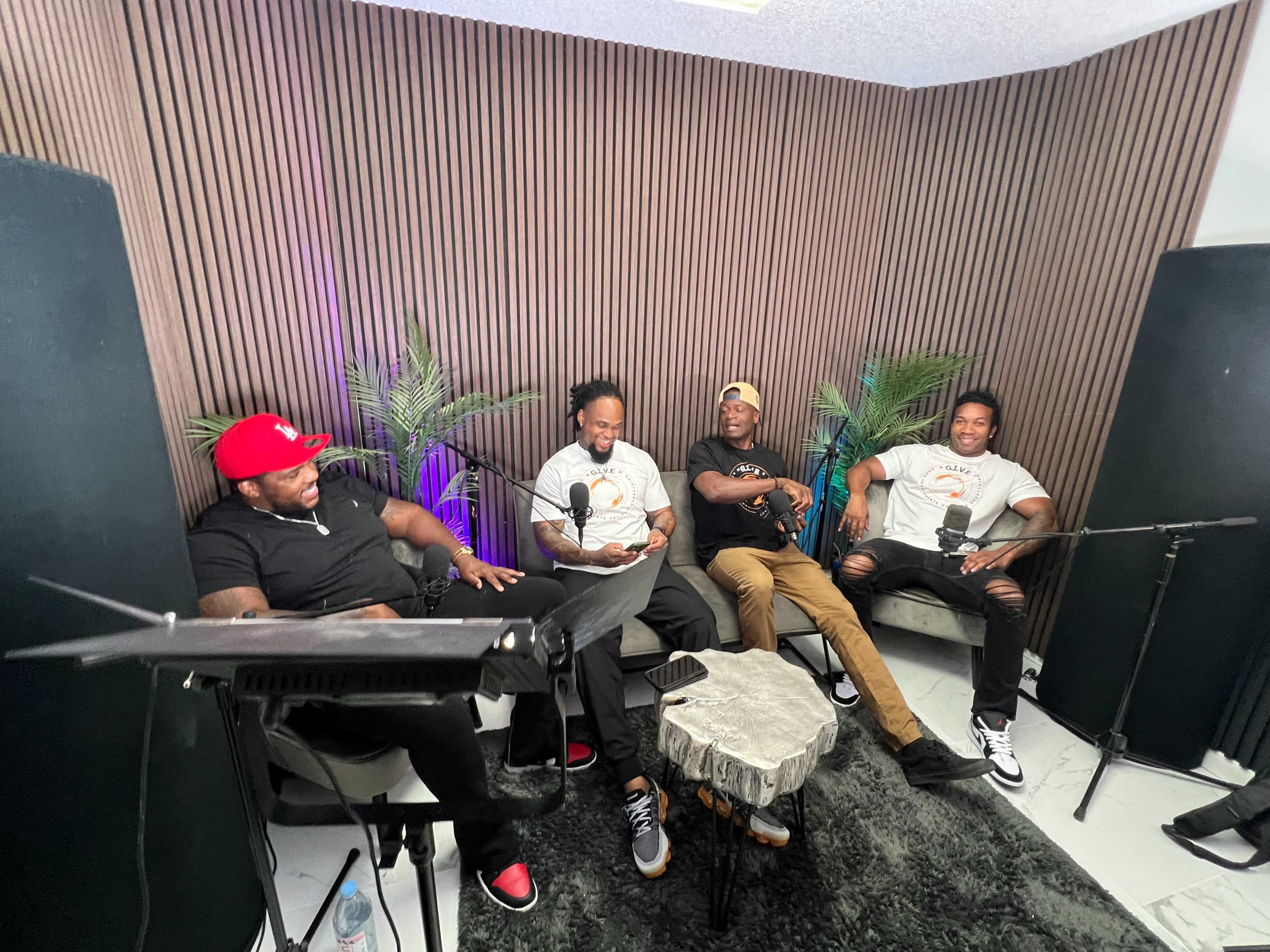 Four men sit in a podcast studio, laughing and conversing while microphones are positioned in front of them.