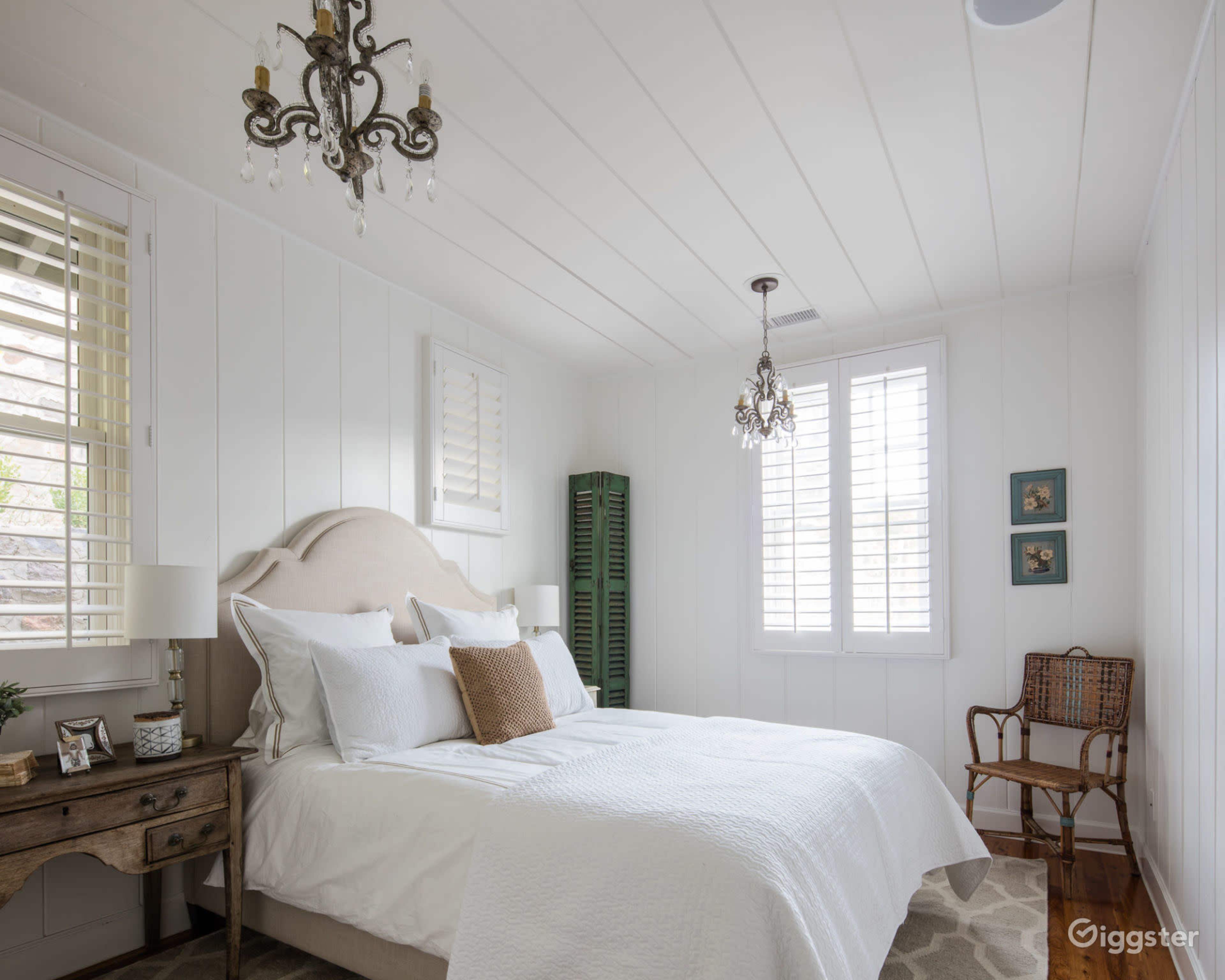 A bright, minimalist bedroom featuring a bed with white linens, a wooden bedside table, a decorative chandelier, and green shutters on the window.