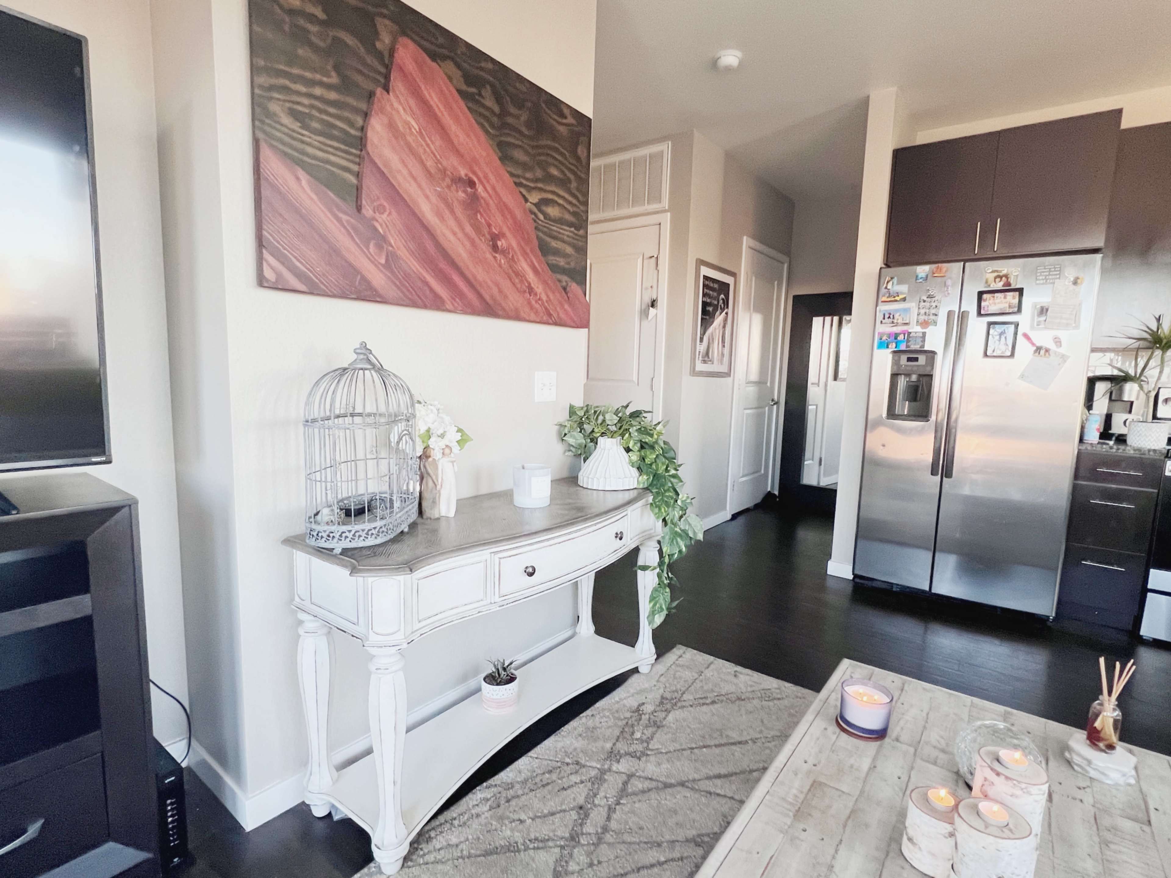 A modern living room with a white console table, a birdcage, and a large wooden art piece on the wall, alongside a stainless steel refrigerator in the background.