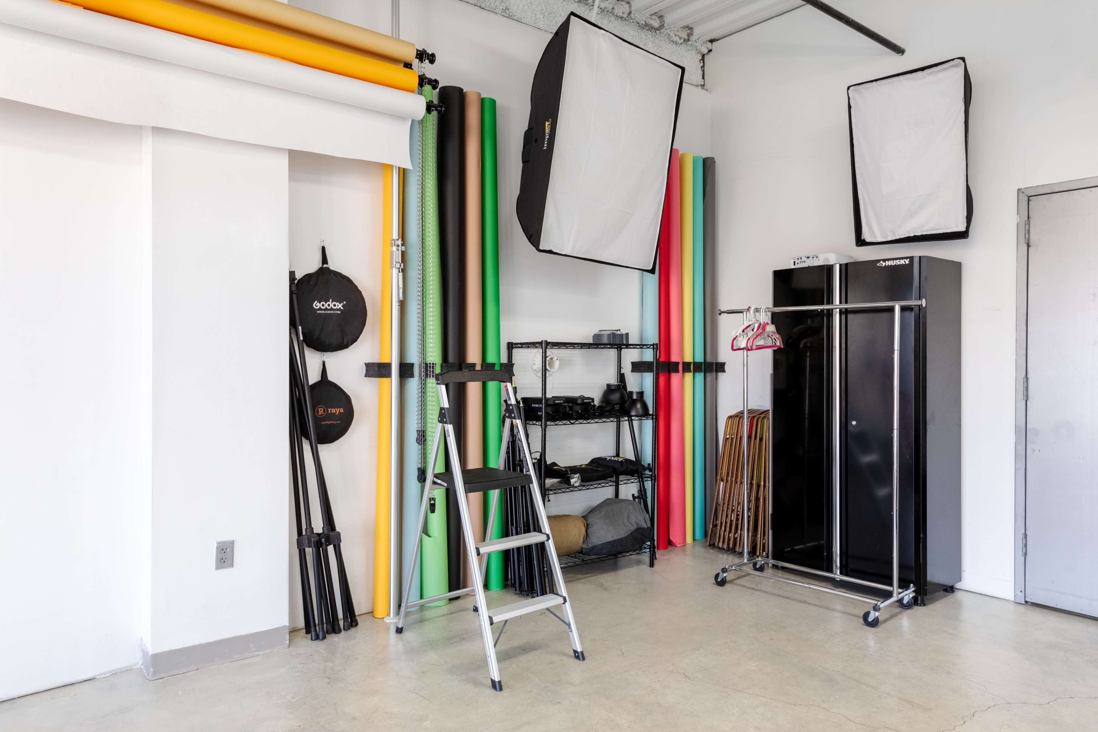 The image shows a photography studio with various colored backdrops, lighting equipment, a ladder, and a clothing rack.