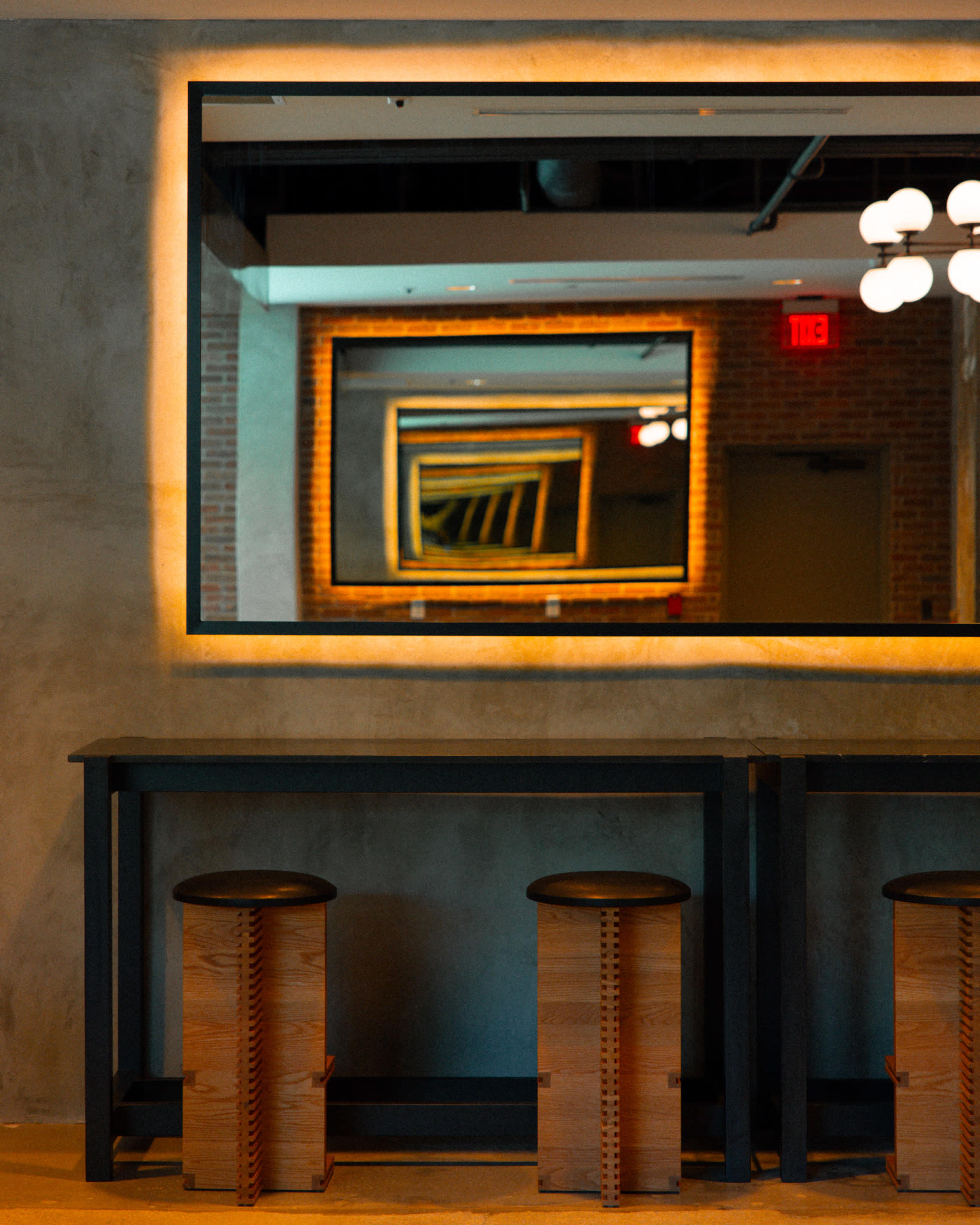 A row of wooden stools sits in front of a large mirror reflecting a series of repeating frames and ambient lighting in a modern interior space.