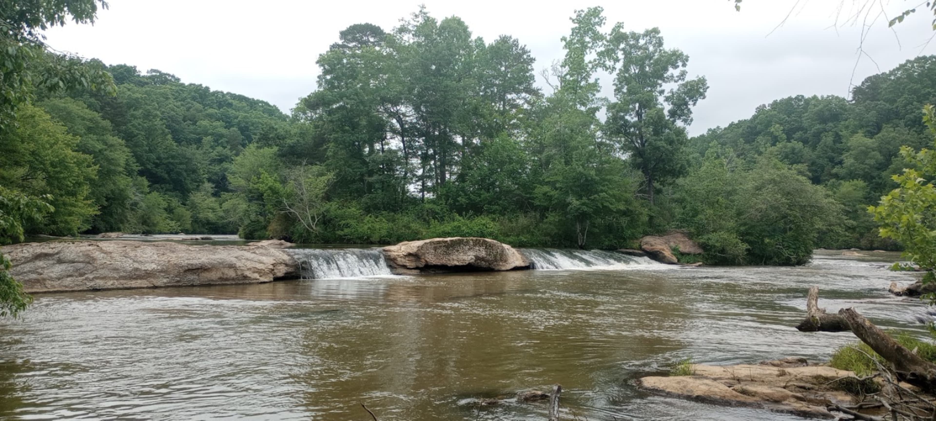 A wide river flows over a rocky ledge, with lush green trees lining the banks and a cloudy sky above.