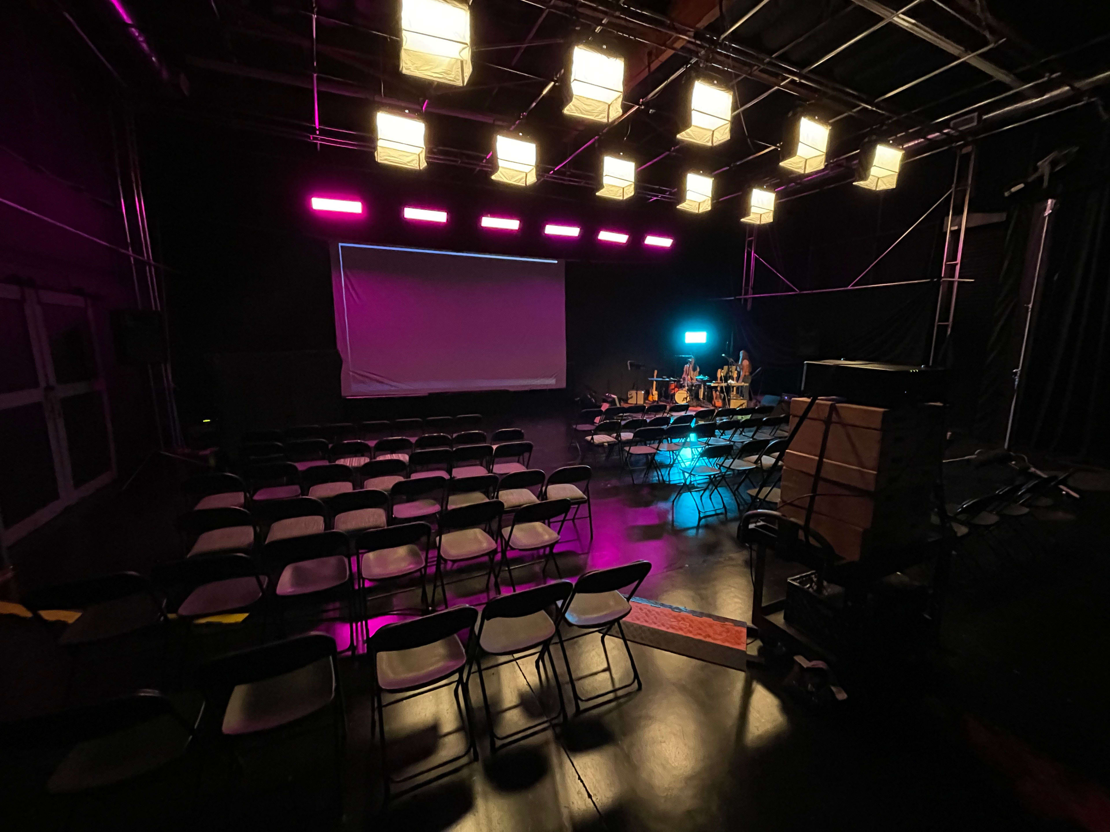A dimly lit stage space features rows of black chairs facing a large screen, with colorful lights illuminating the area.