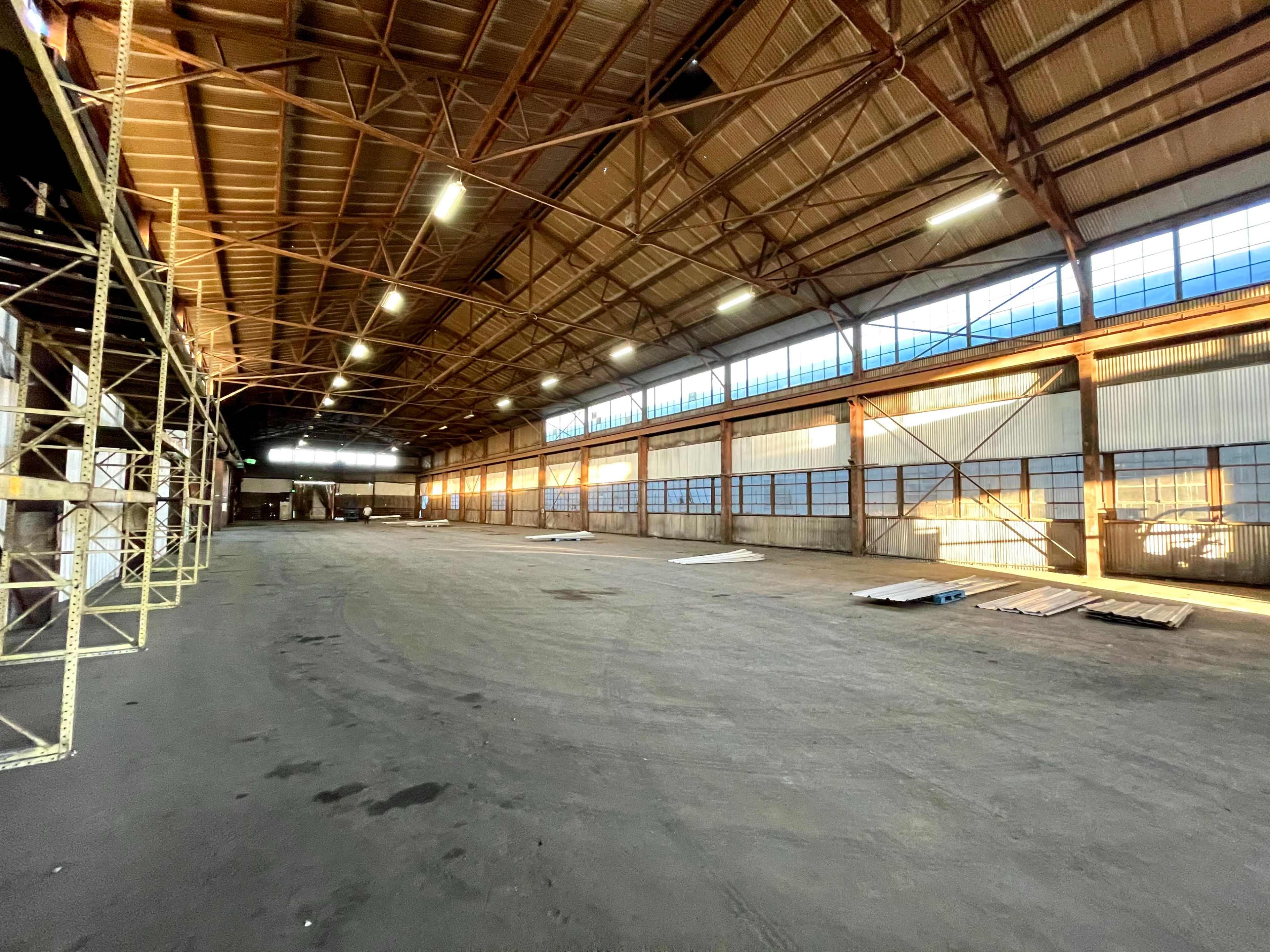 A large, empty industrial warehouse with metal beams and scattered materials on the floor.