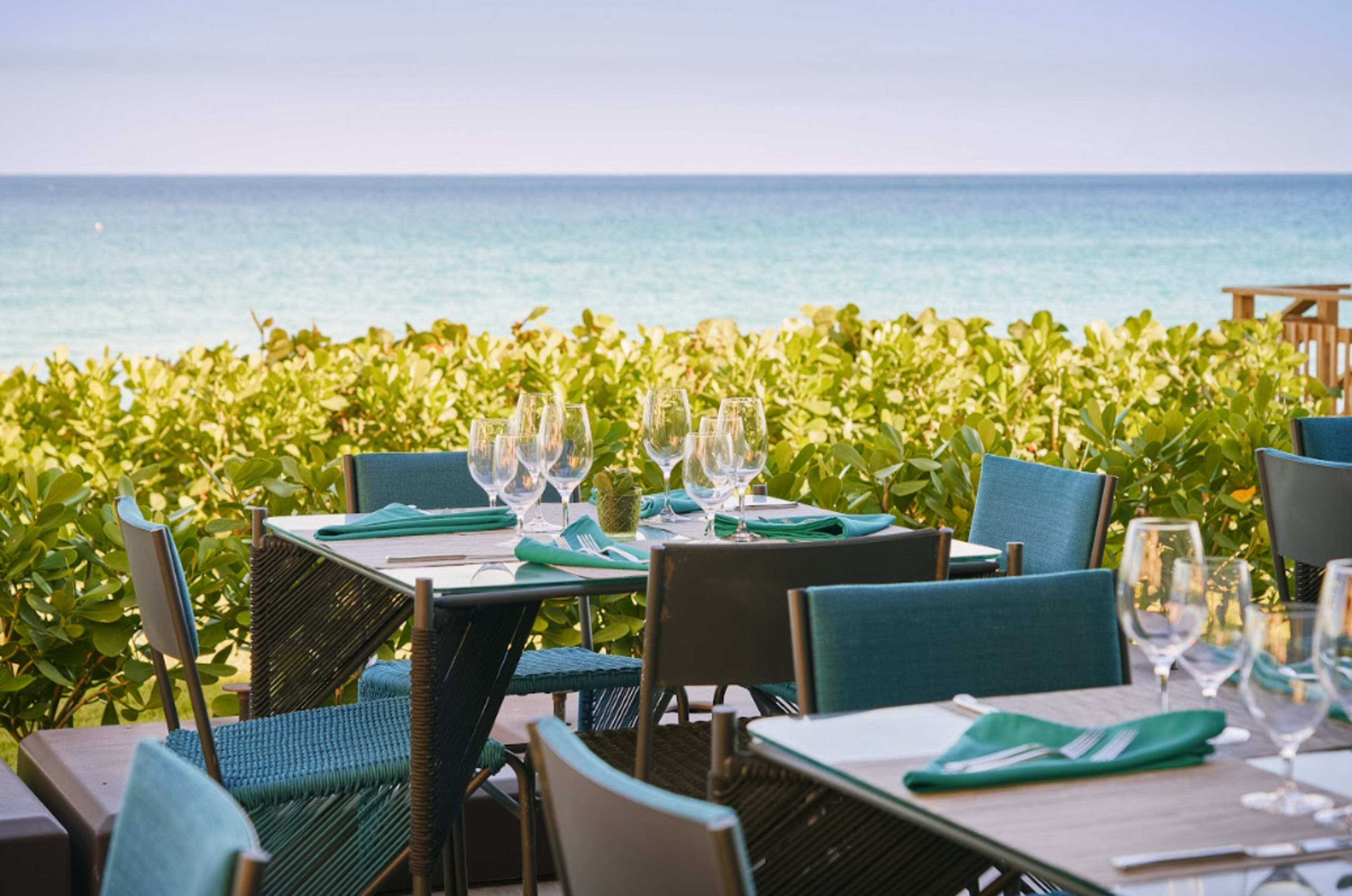 A seaside restaurant features tables set with glassware and turquoise accents, overlooking a calm ocean and lush greenery.