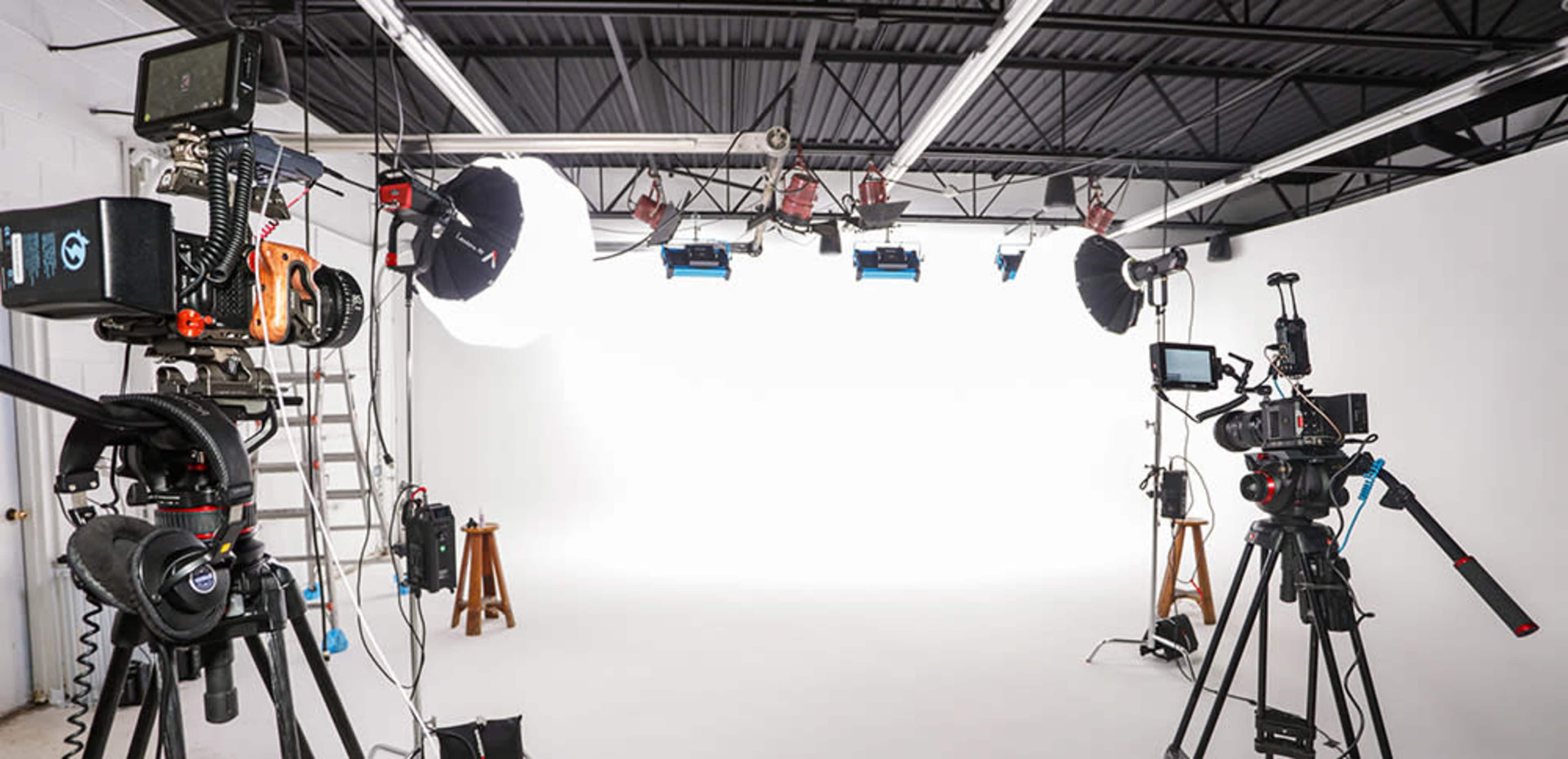 A photography studio setup with multiple cameras, lighting equipment, and a blank white backdrop.