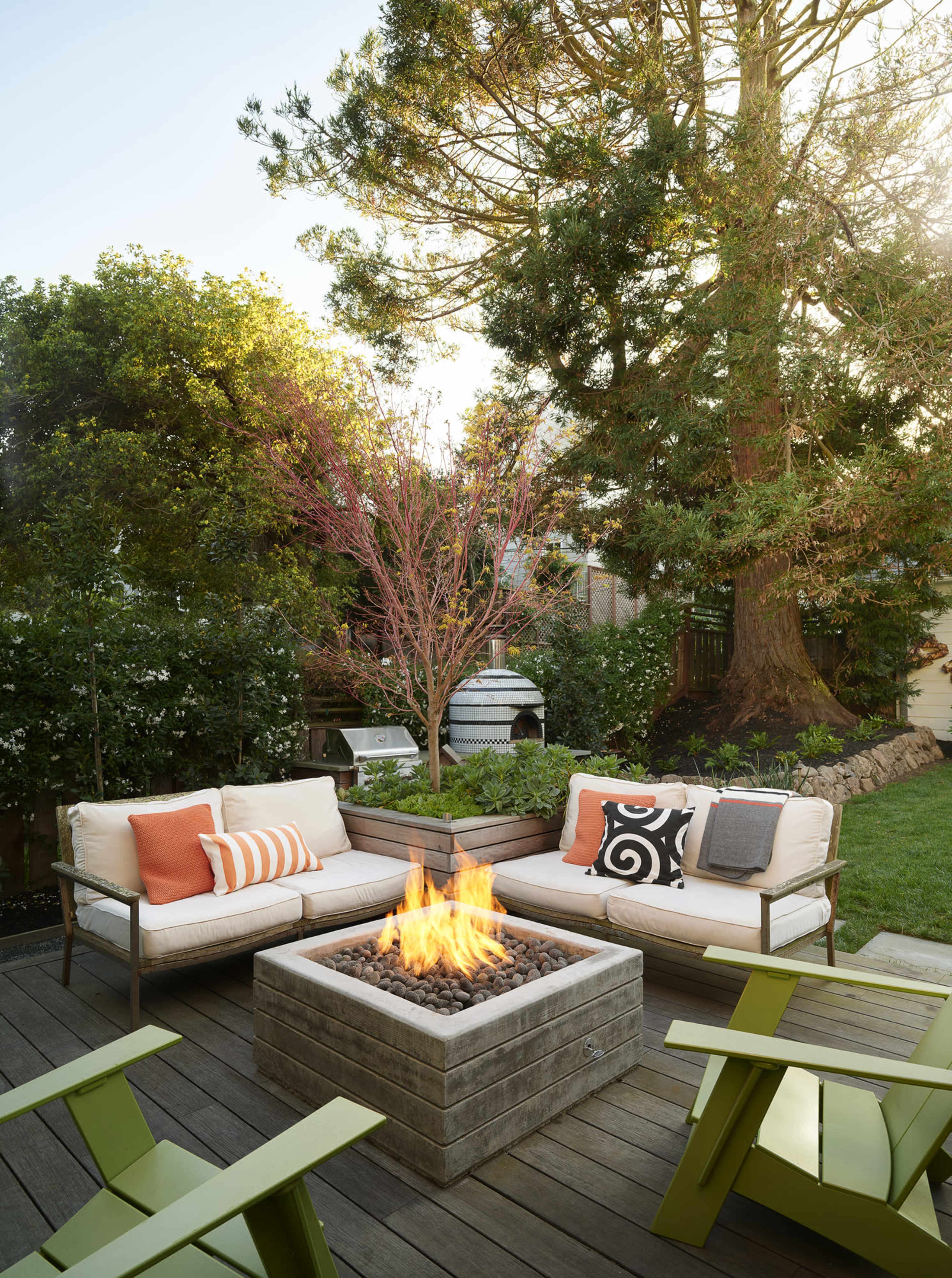 A cozy outdoor seating area features a fire pit surrounded by comfortable couches and vibrant green chairs, set against a backdrop of trees and garden greenery.