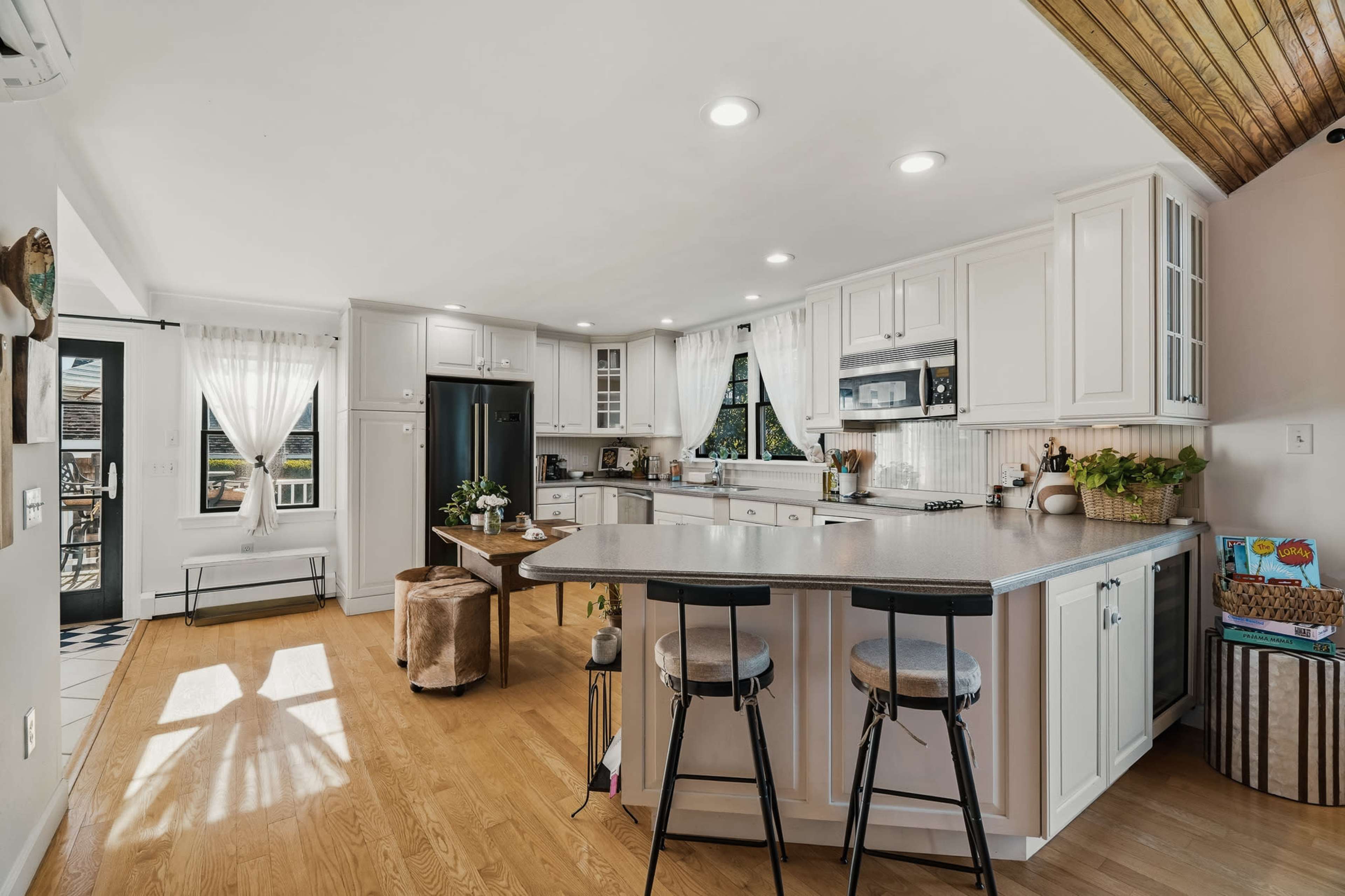 A modern kitchen features a central island with bar stools, white cabinetry, and large windows providing natural light.