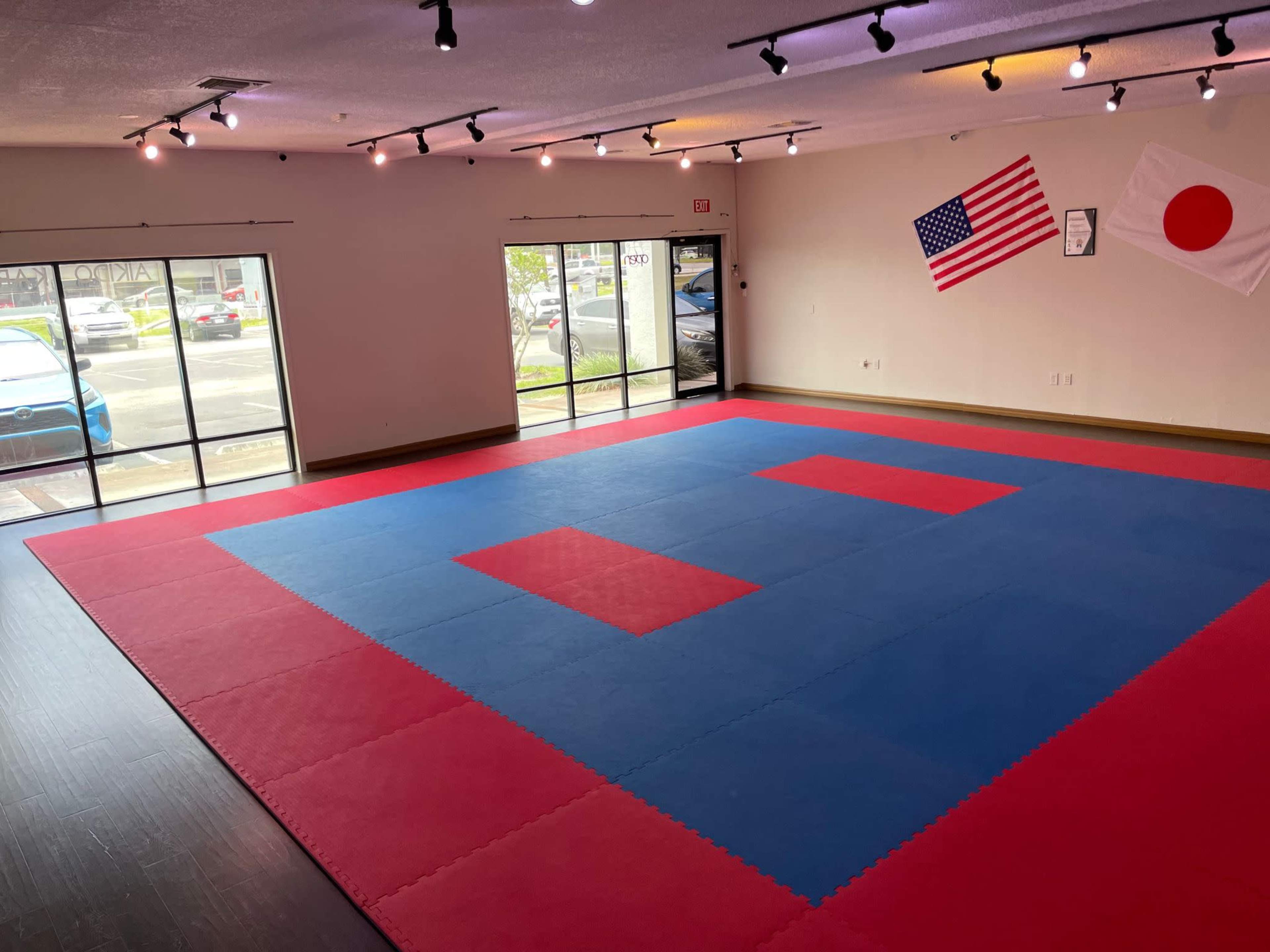 An empty martial arts dojo with red and blue mats and flags of the United States and Japan hanging on the walls.