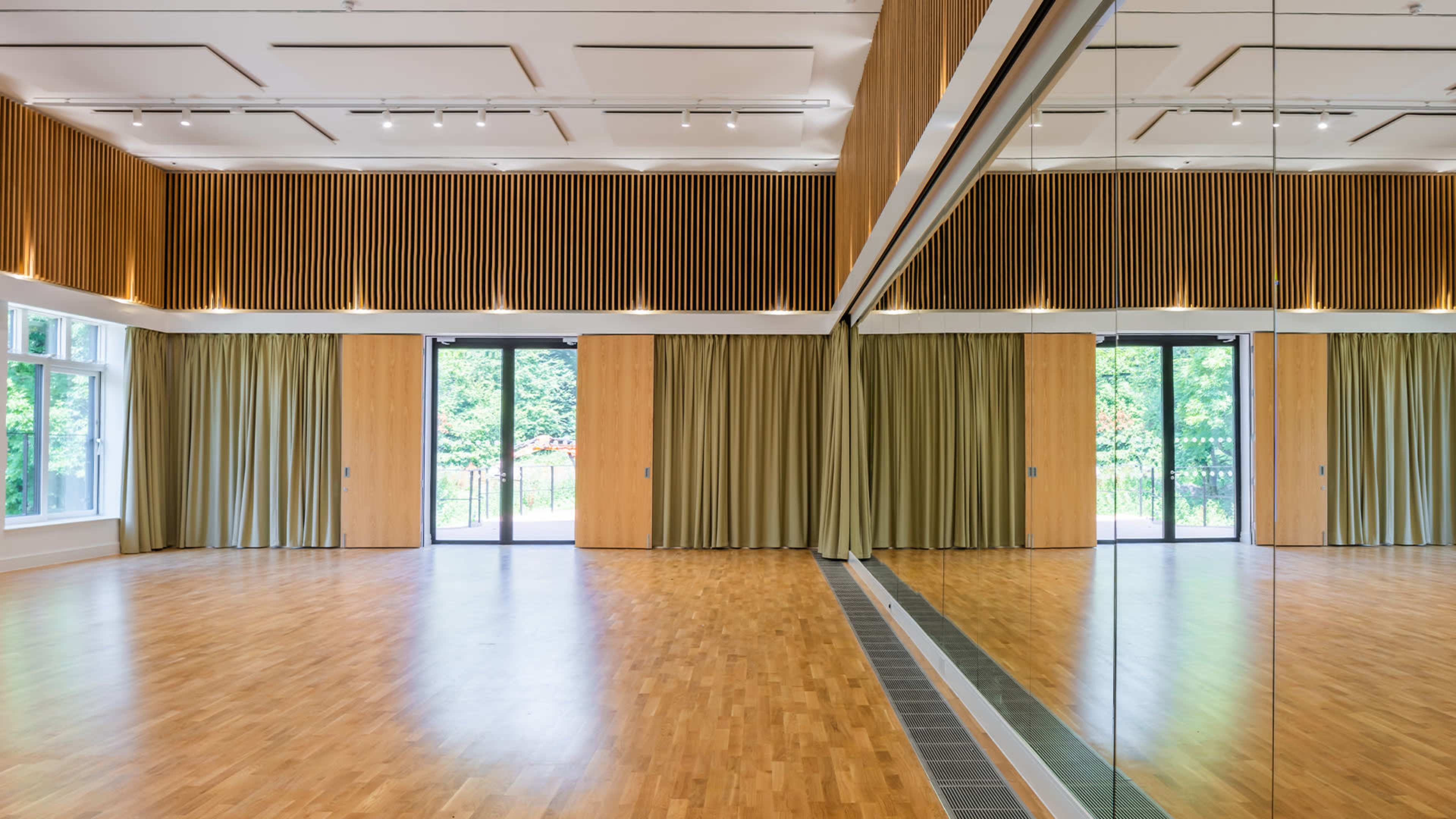 A spacious, wooden-floored room with large mirrors on one wall and floor-to-ceiling curtains on either side of a pair of glass doors leading to an outdoor area.