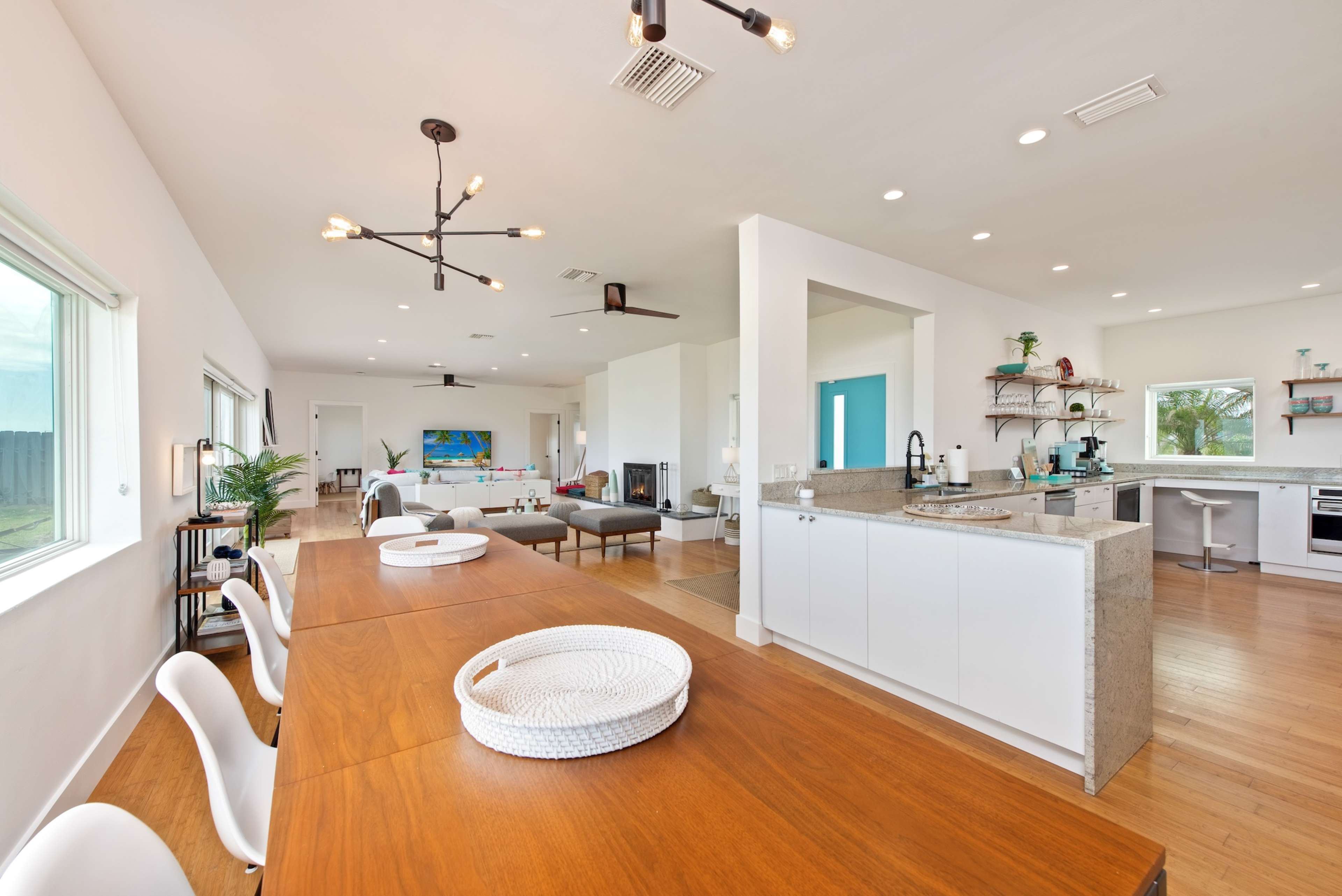 The image shows a spacious modern interior with a dining area leading into an open kitchen and living space, featuring large windows and light-colored walls.
