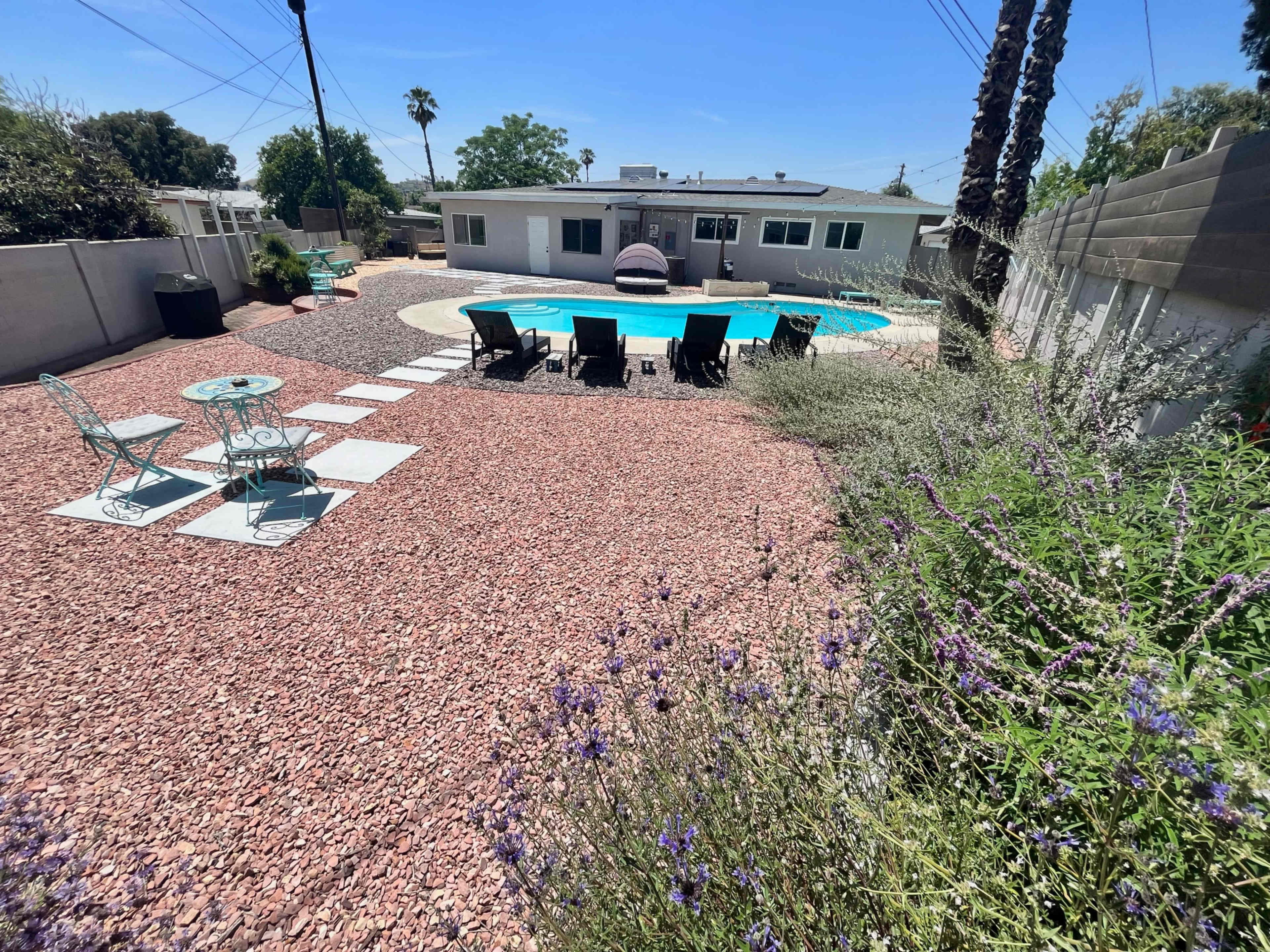 A backyard featuring a swimming pool surrounded by a gravel landscape, patio furniture, and several plants.