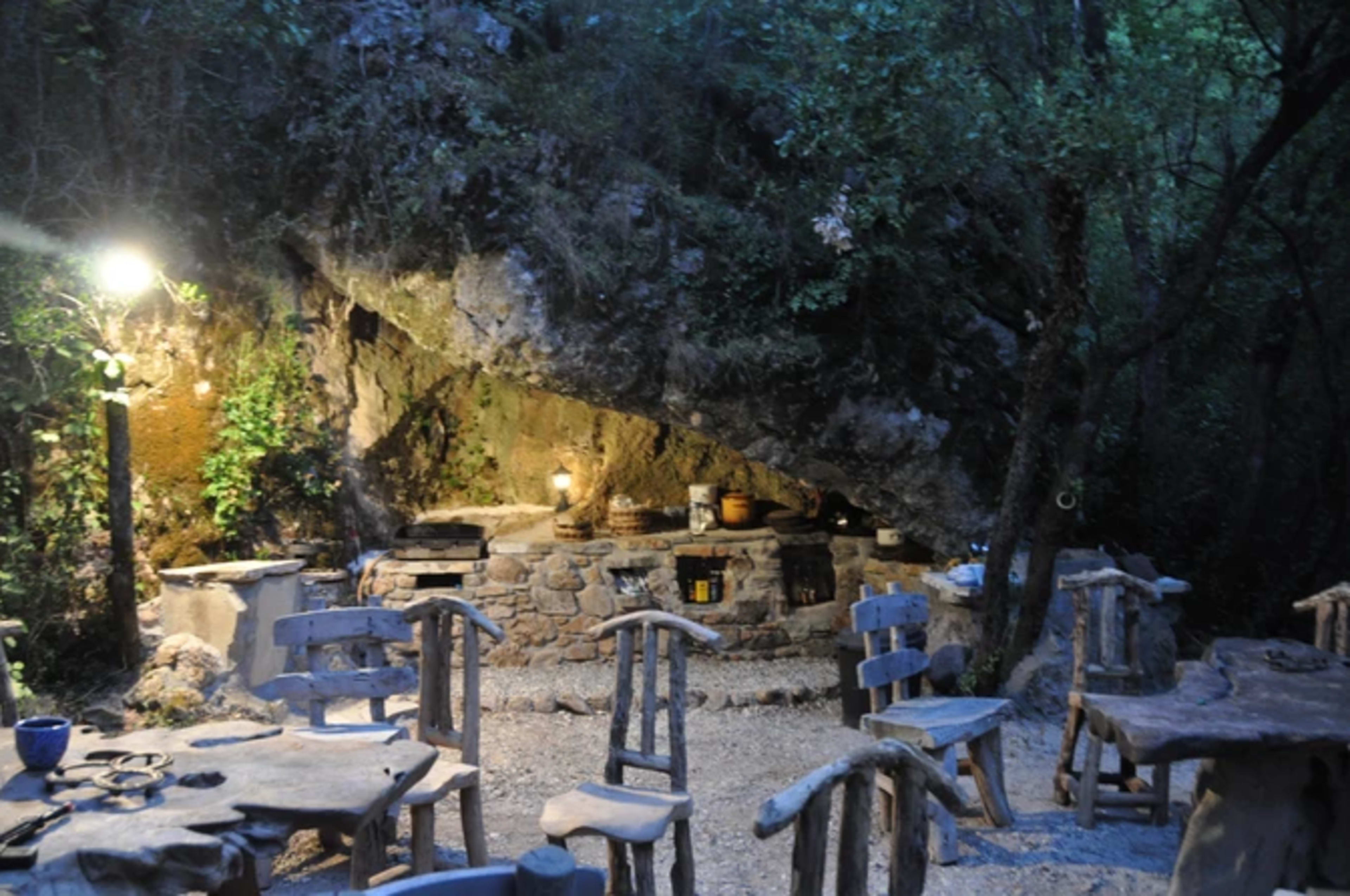 The image shows an outdoor dining area with stone tables and chairs under a rocky overhang surrounded by trees, illuminated by a lamp.