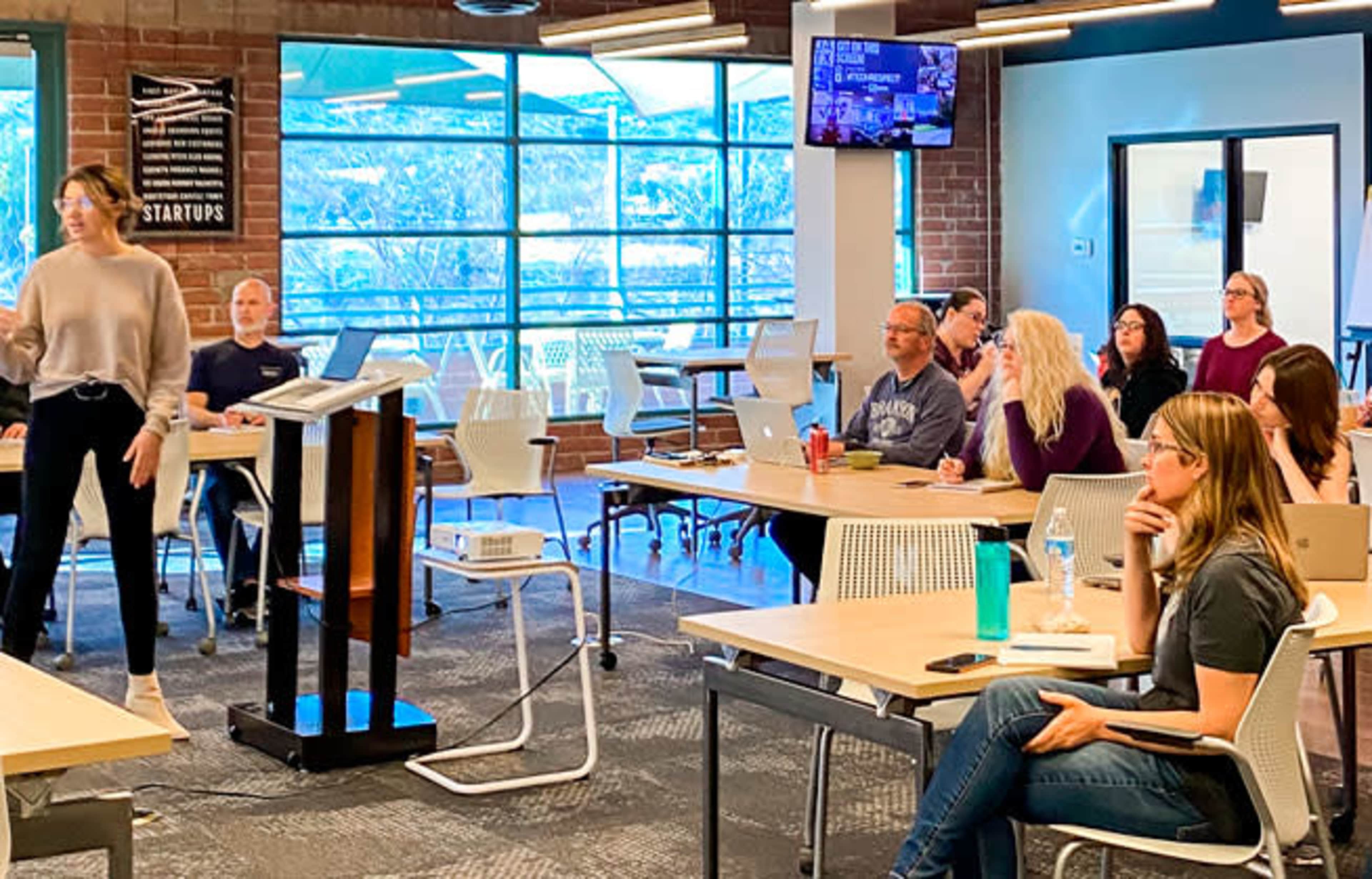 A presenter stands in front of a group of seated attendees in a modern conference room with large windows and a presentation screen.