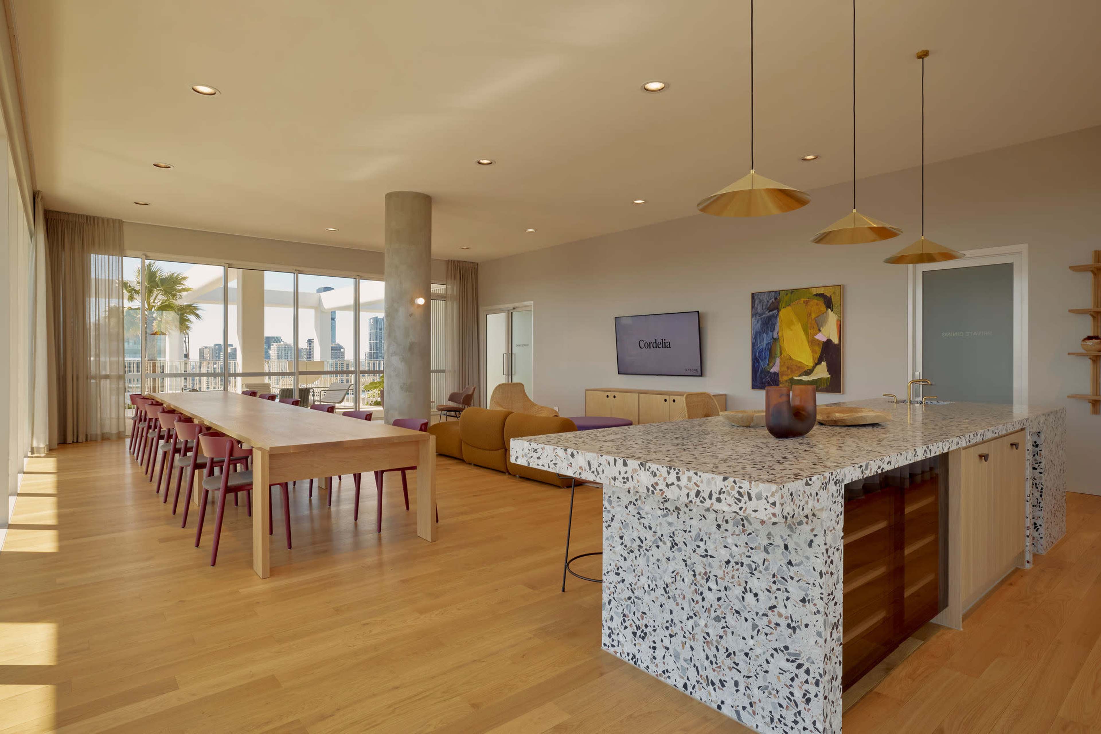 The image shows a modern open-concept living area with a large dining table, an island bar in the kitchen, and floor-to-ceiling windows offering views of a city skyline.