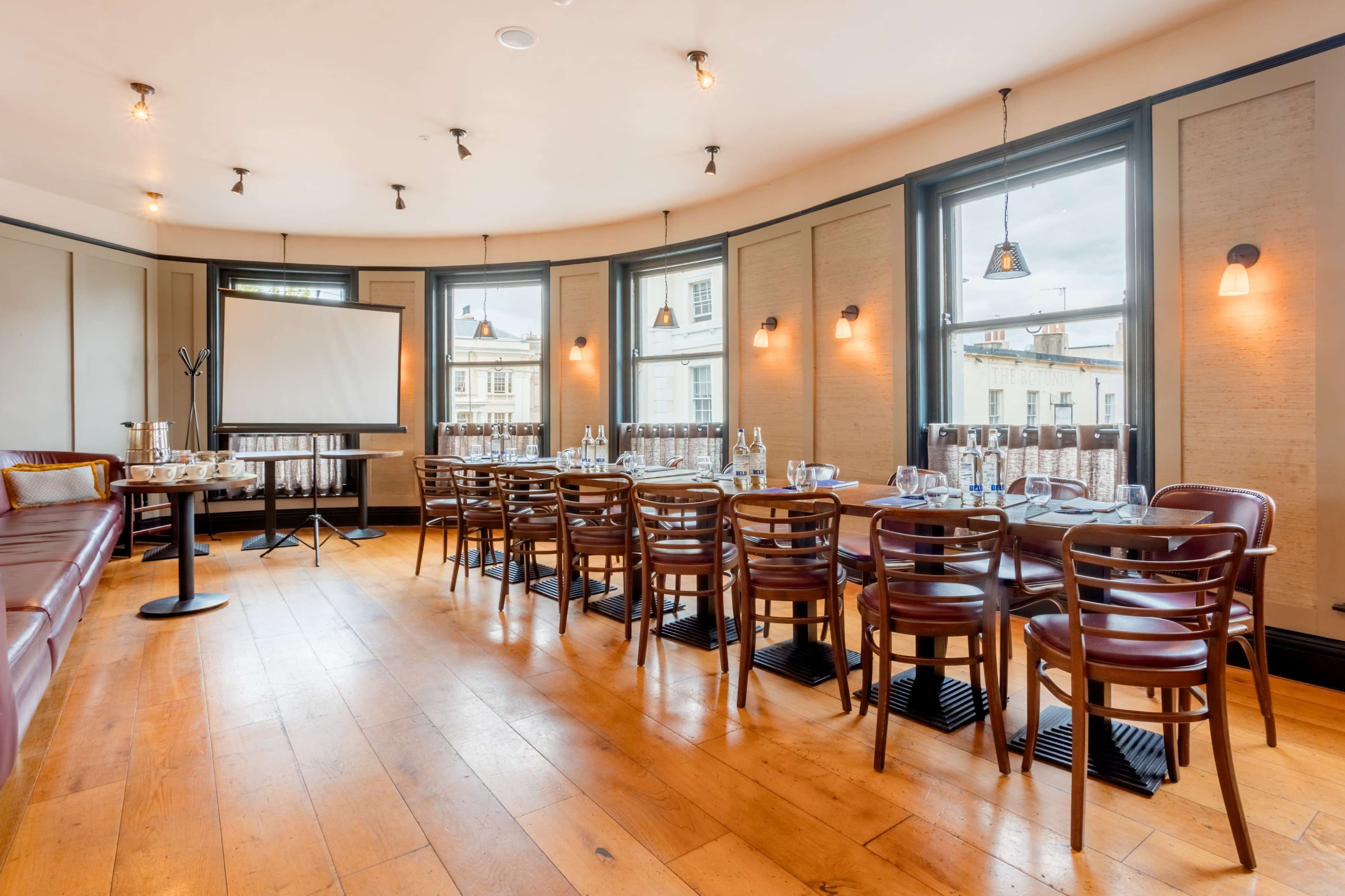 The image shows a dining area with multiple wooden tables arranged in a U-shape, surrounded by chairs, and featuring large windows that provide natural light.