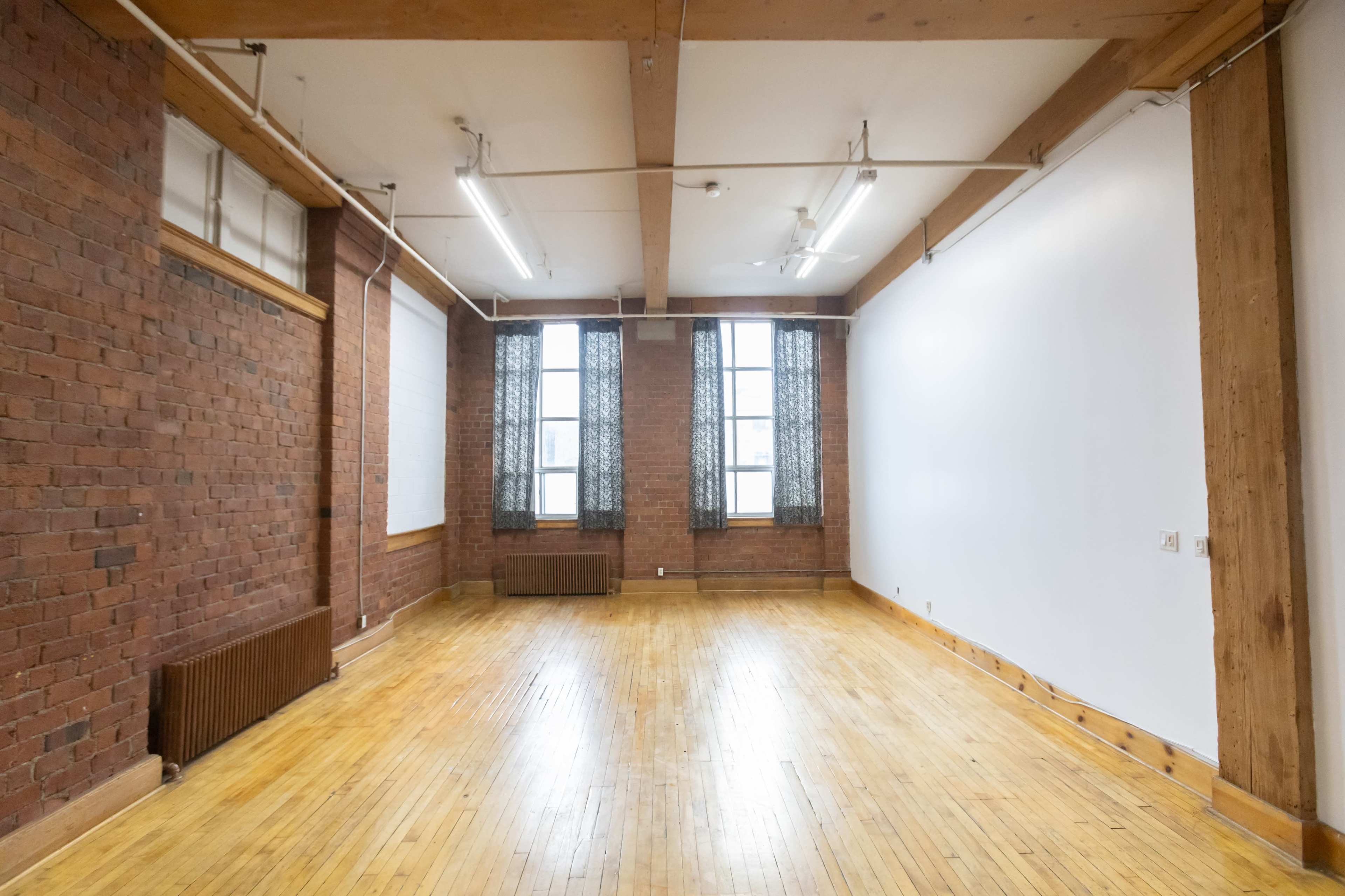The image shows a spacious room with exposed brick walls, large windows, and polished wooden floors.