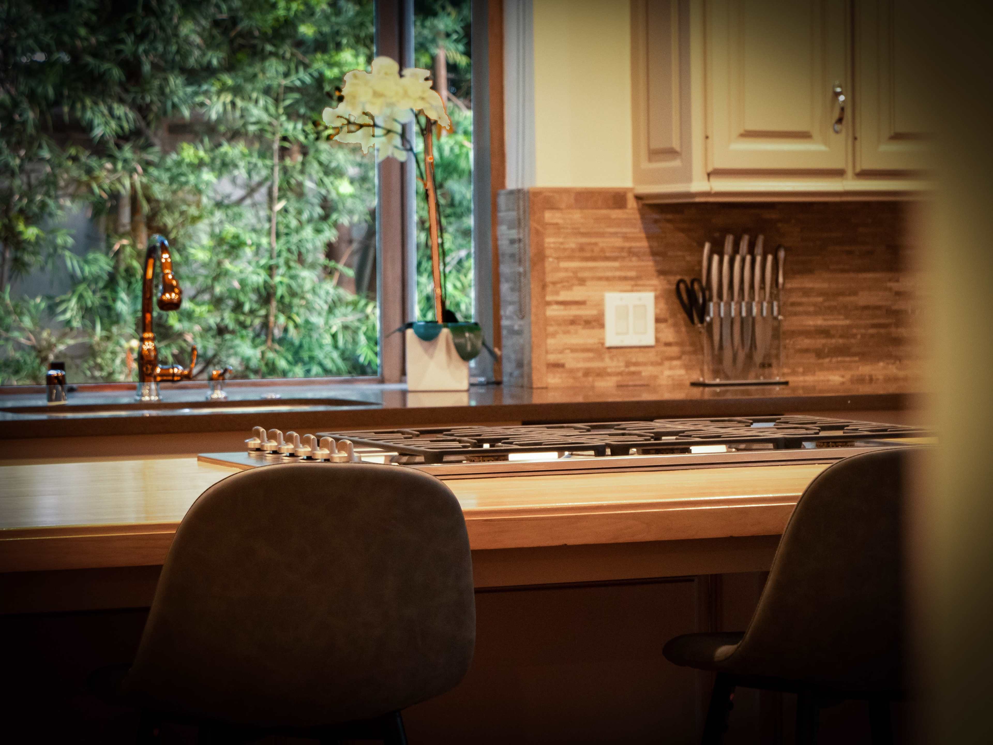 A modern kitchen features a central island with a stovetop, two chairs, and a window overlooking greenery.