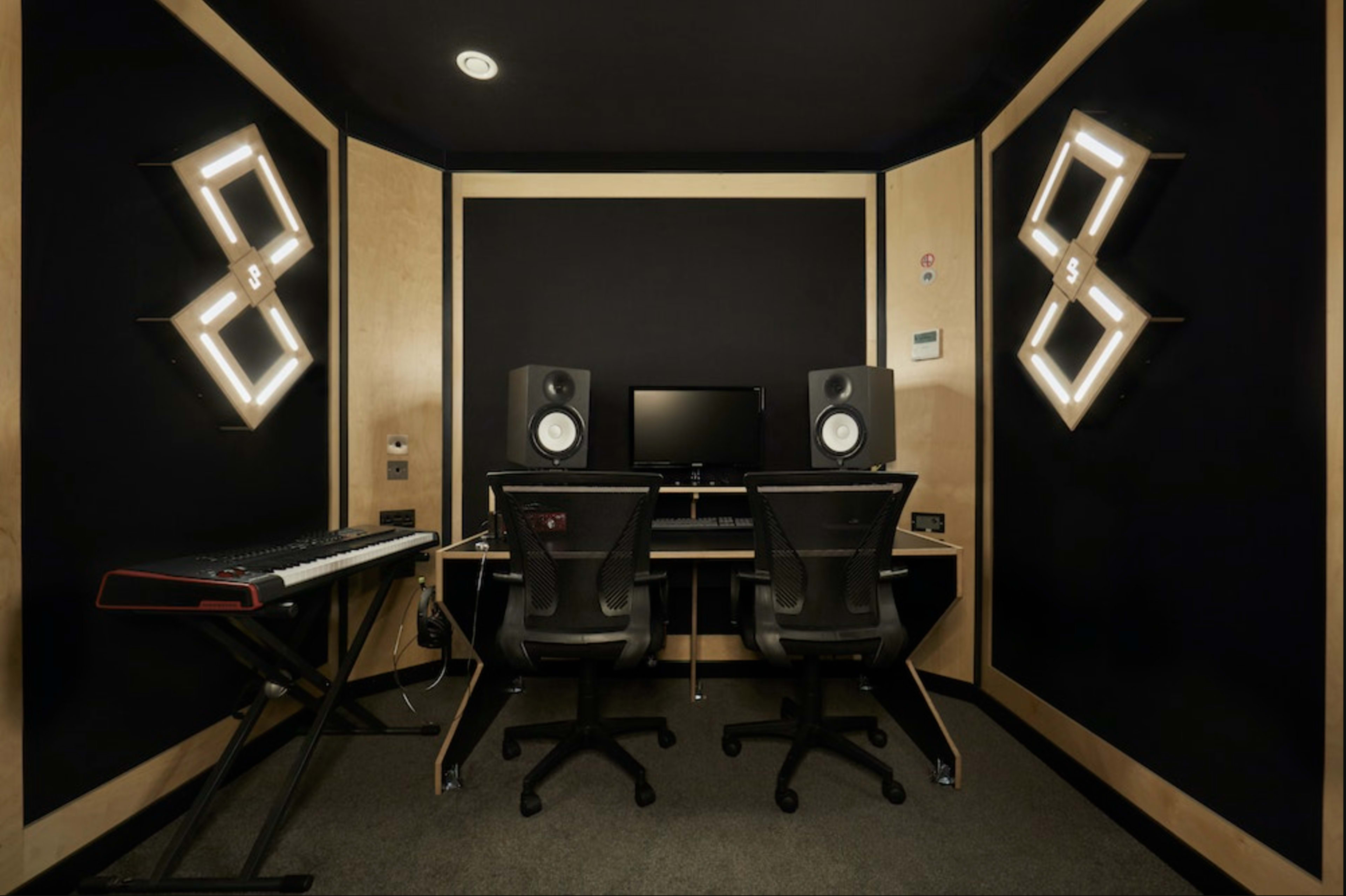 A music production studio with two black chairs, a computer monitor, speakers, and a keyboard in a small, dark room.