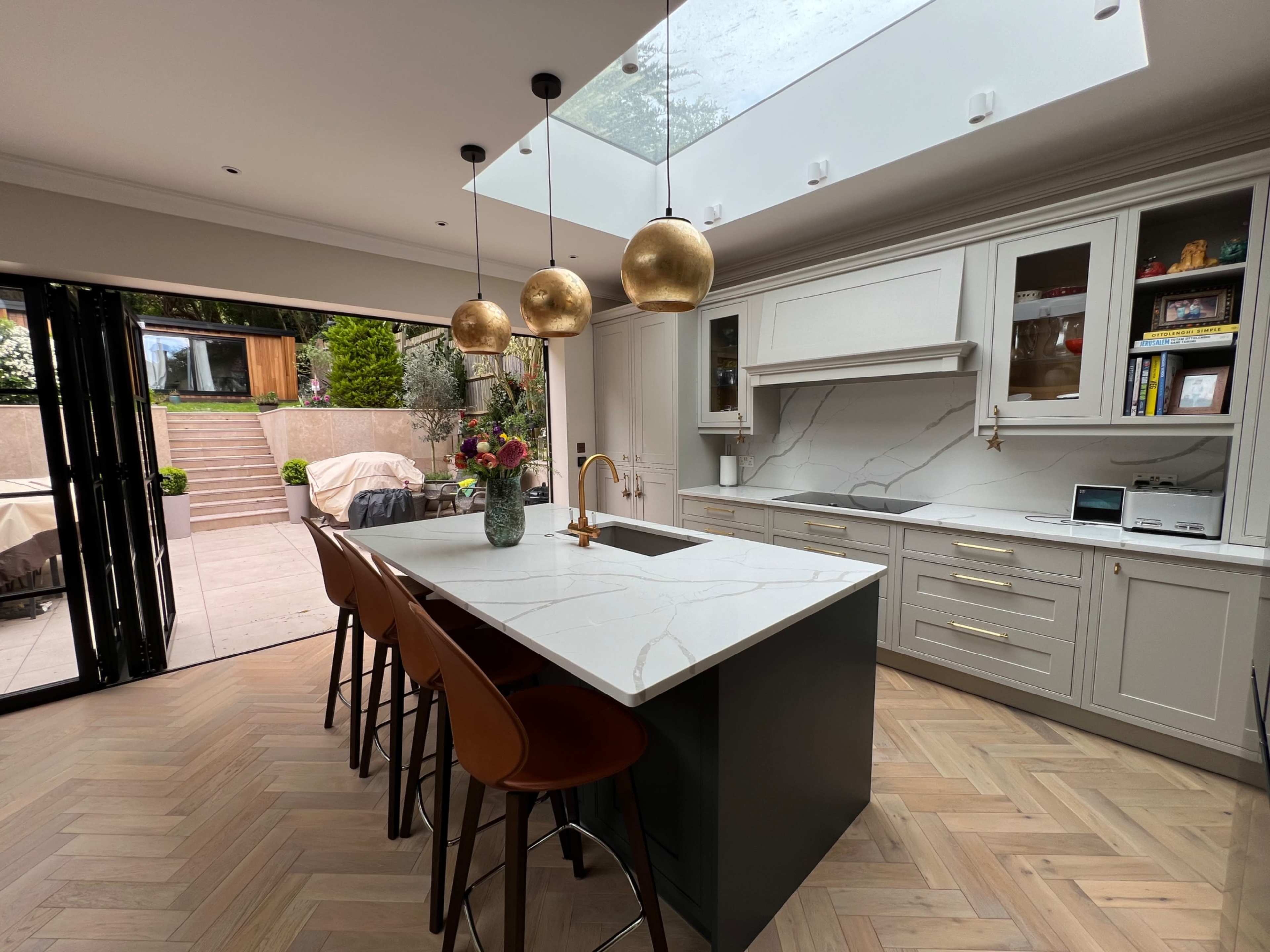 A modern kitchen features a central island with bar stools, pendant lighting, and a large skylight above, with sliding doors that open to an outdoor space.