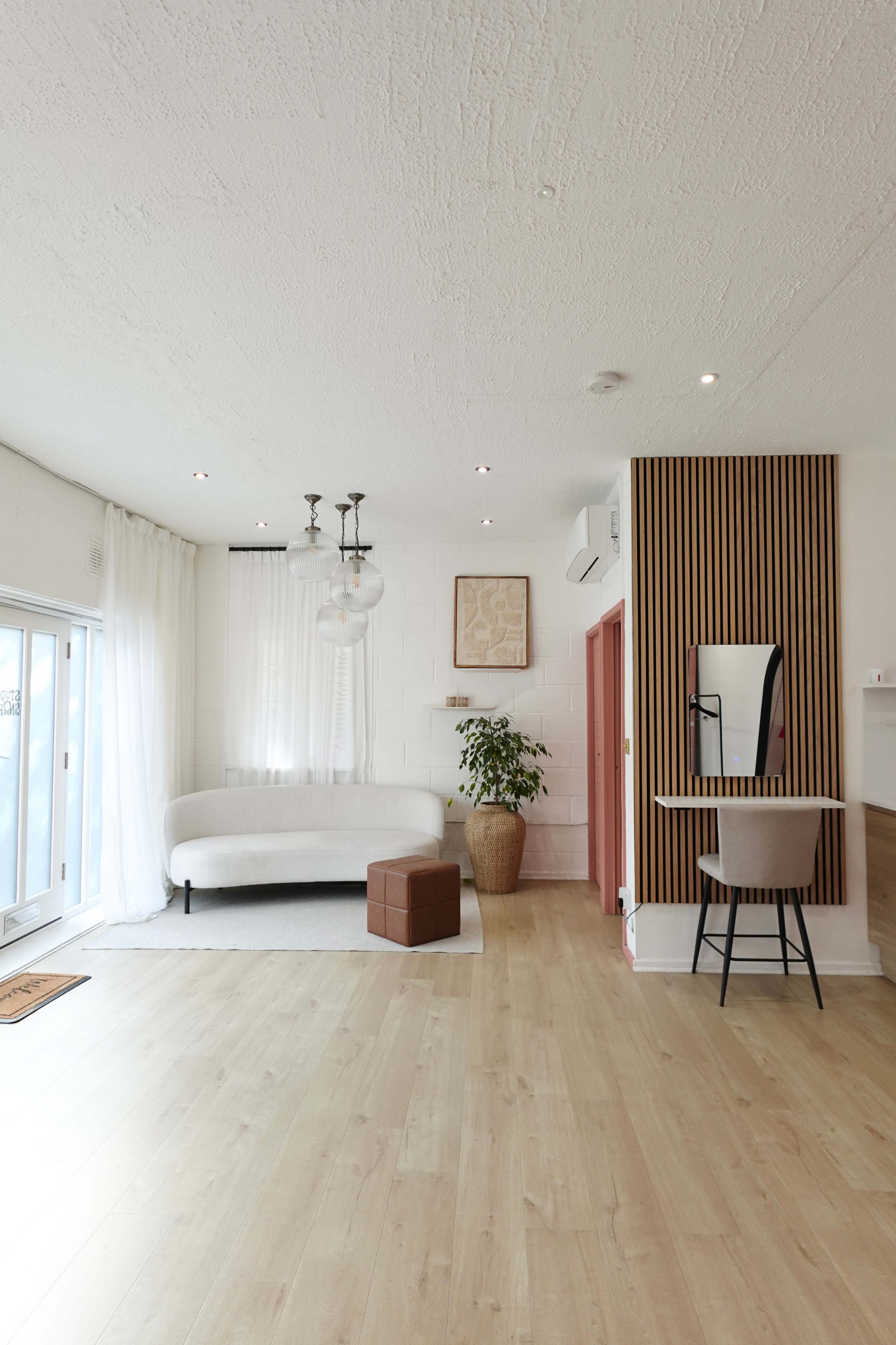 The image shows a bright, modern living space with a white curved sofa, large windows, a potted plant, and a small brown box in the center.