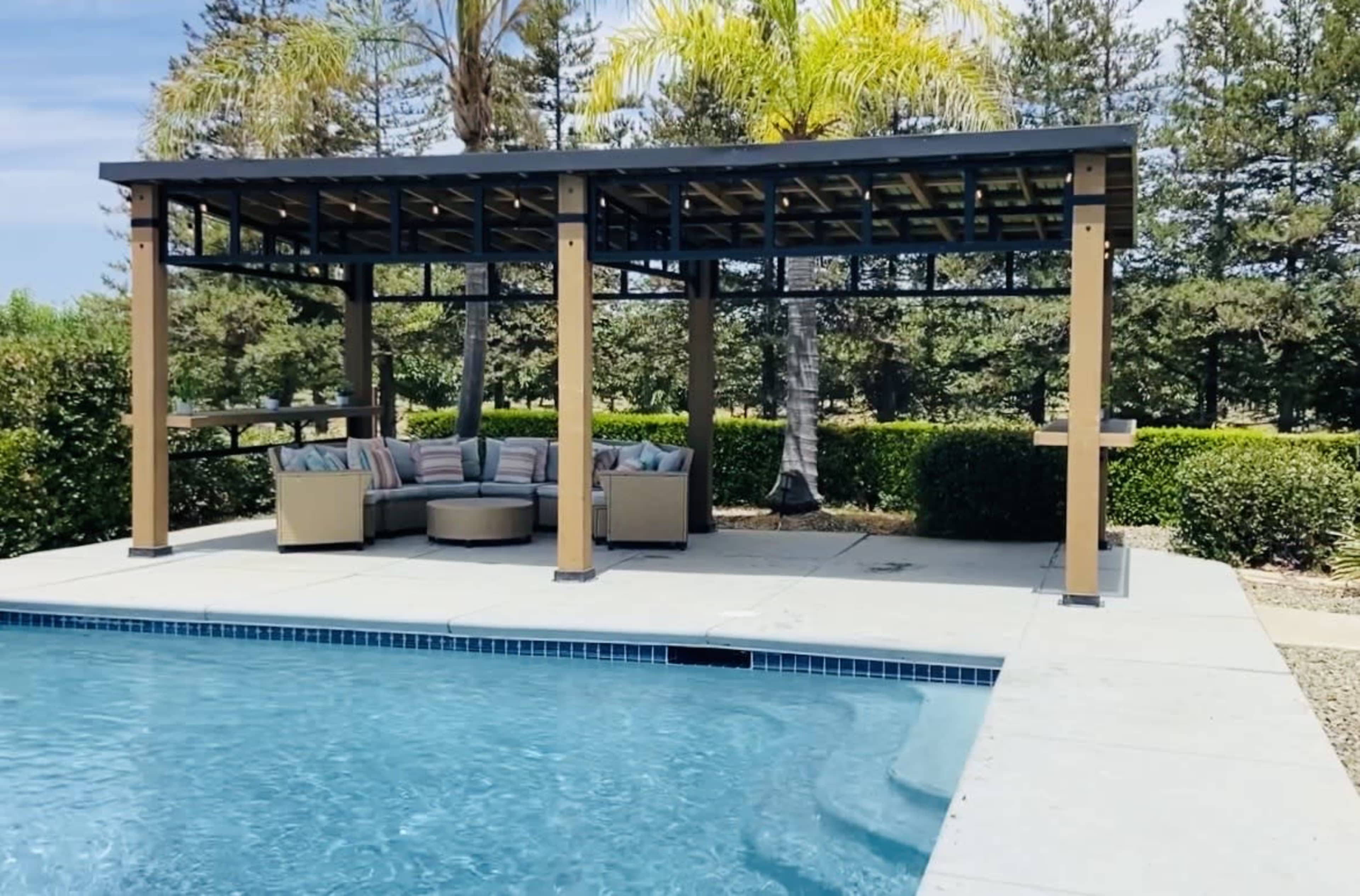 A shaded seating area with cushions is located near a swimming pool surrounded by greenery.