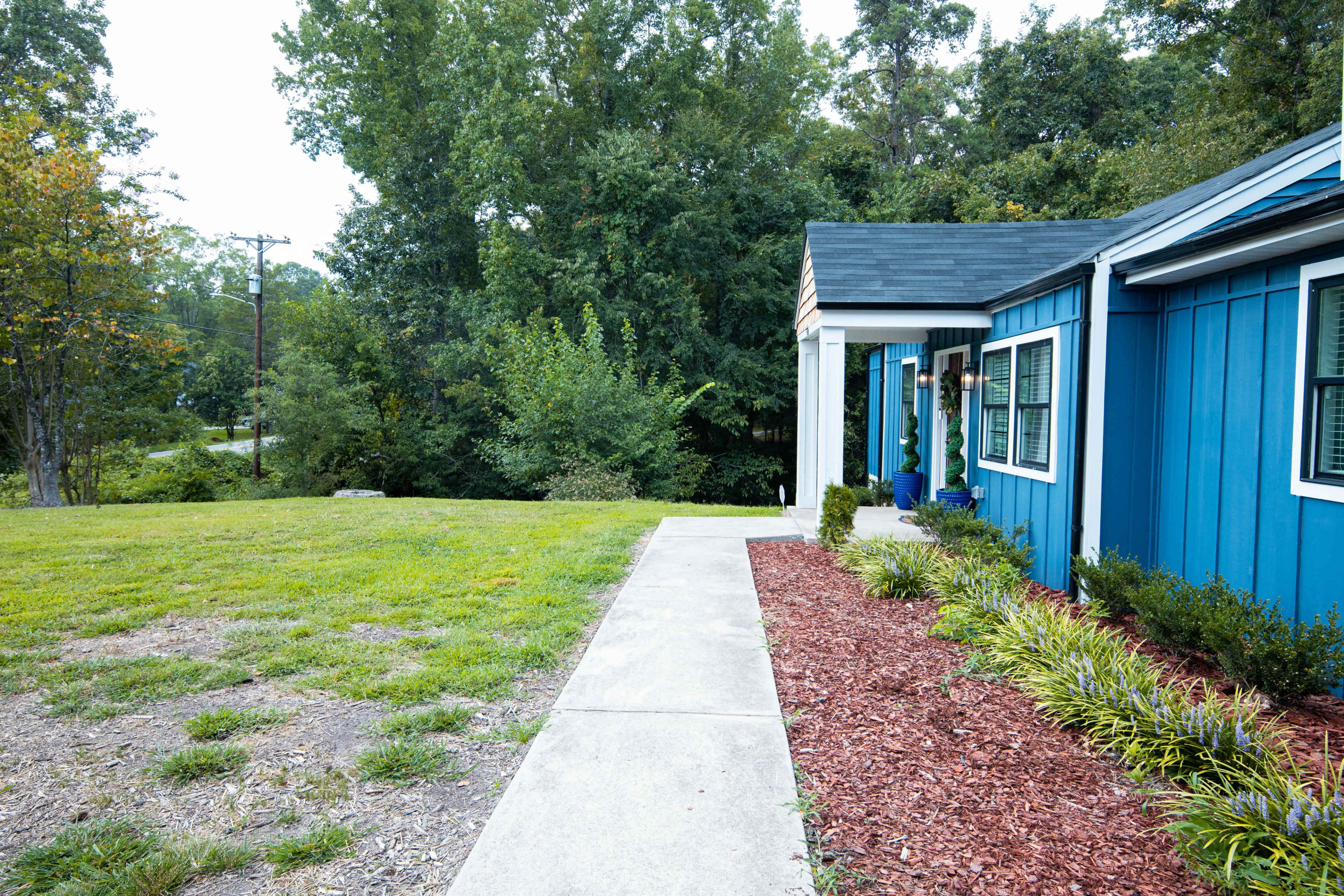 A blue house with a walkway leads to a grassy area and trees in the background.