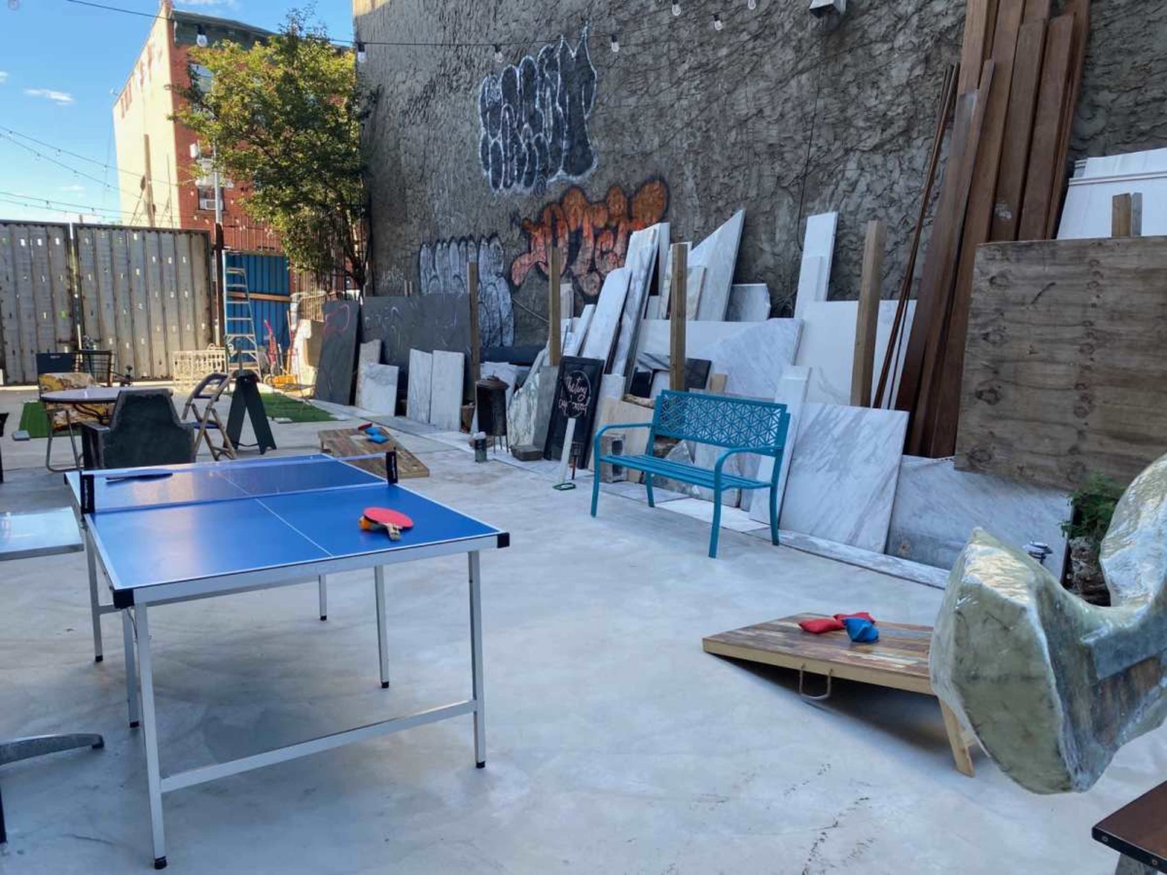 The image shows an outdoor area with a blue ping pong table, various construction materials, and scattered furniture against a graffiti-covered wall.