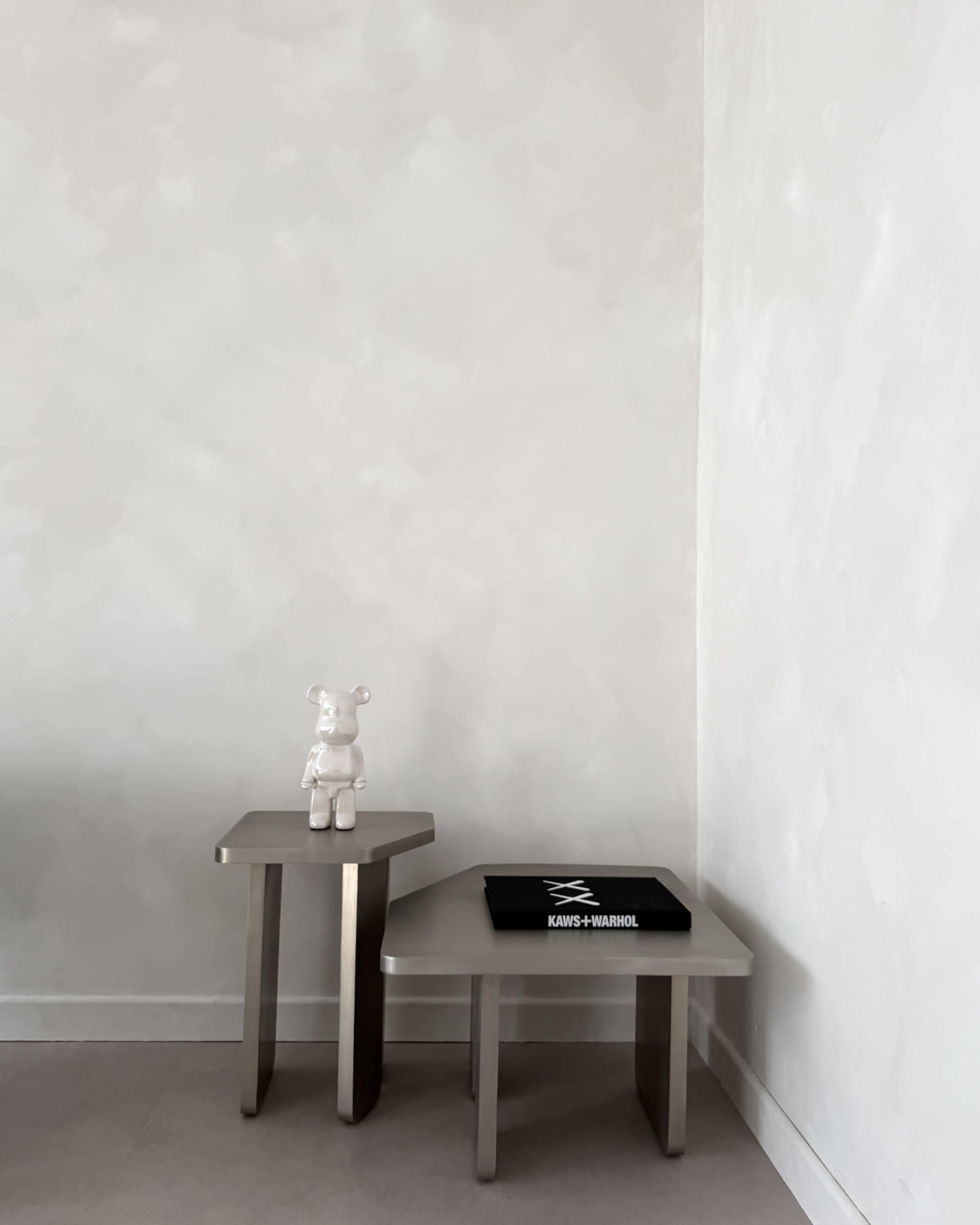 Two minimalist side tables are positioned against a plain, light-colored wall, with a figurine on one table and a black book on the other.