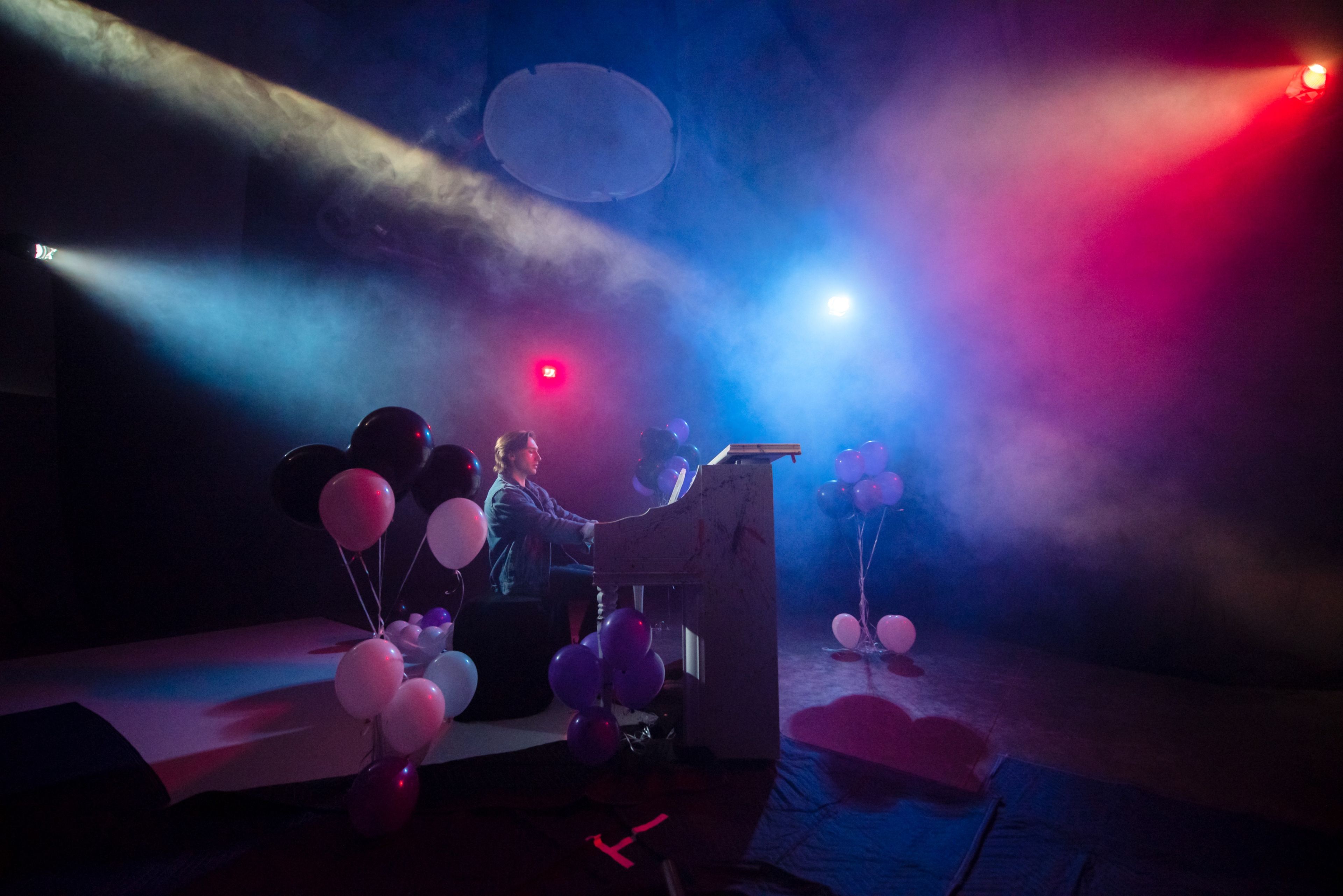 A musician plays a piano on a stage decorated with black, white, and purple balloons, surrounded by colored lighting and fog.