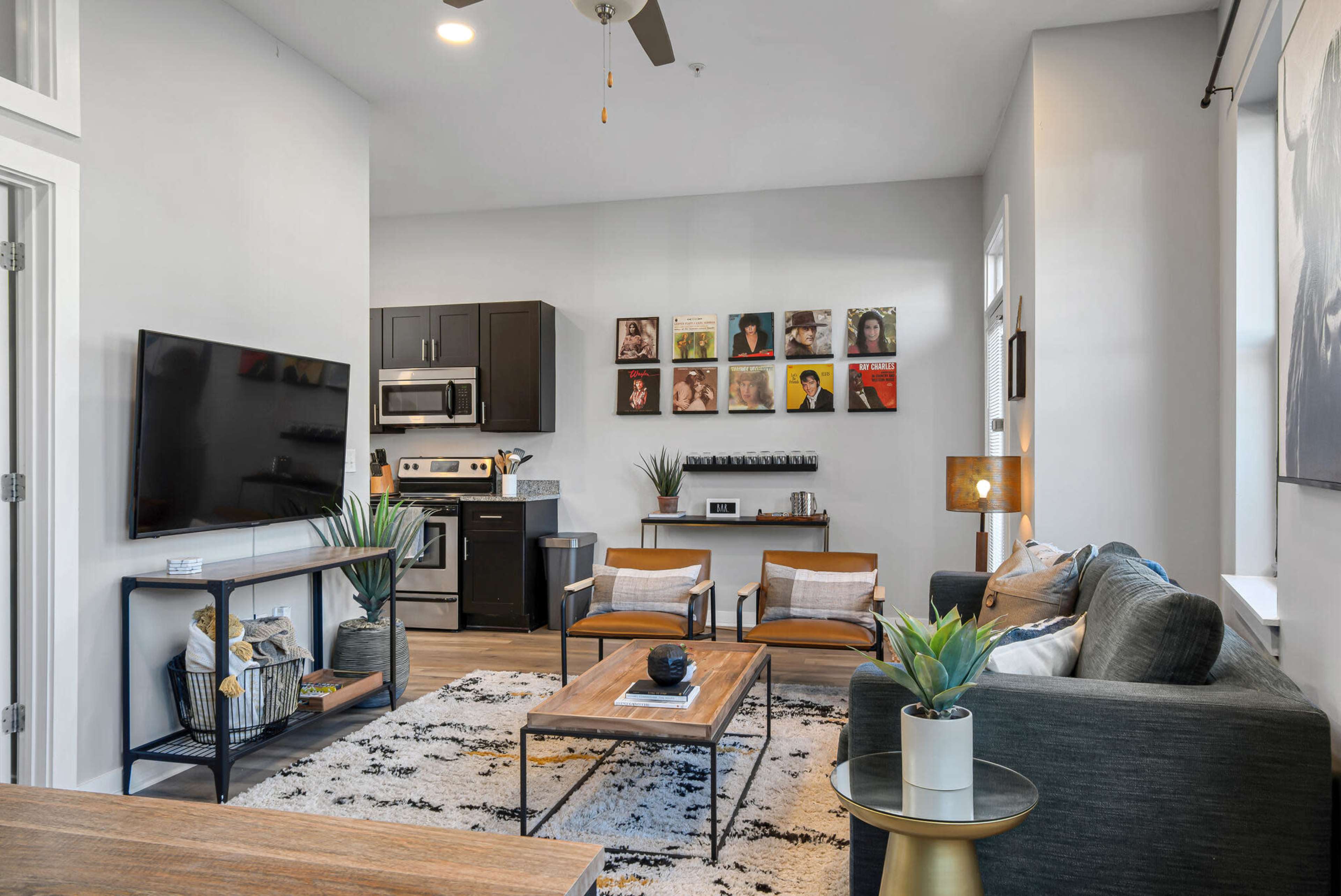 The living room features a modern design with a television, a kitchen area, two armchairs, and a coffee table, surrounded by decorative plants.