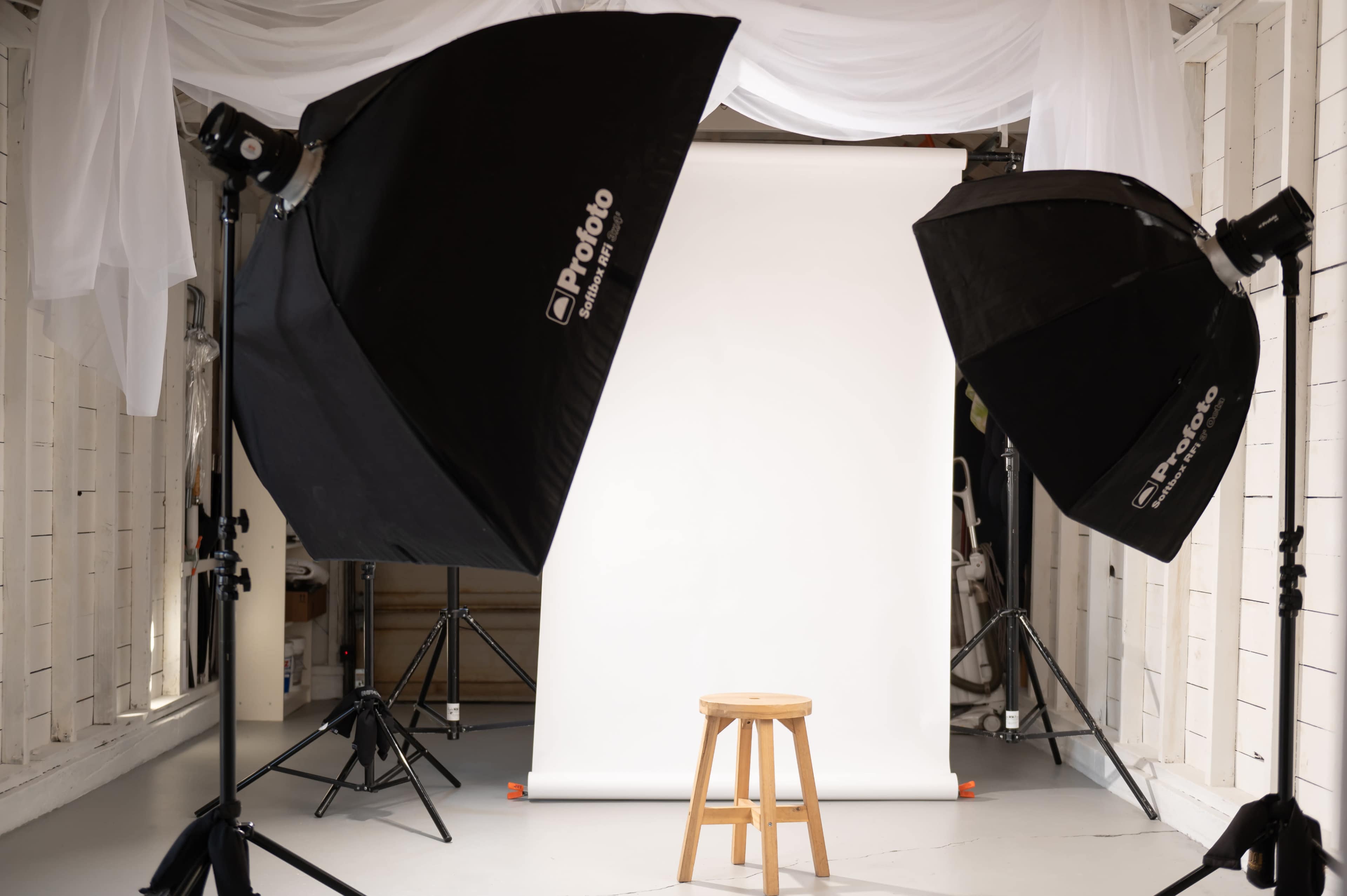 A photography studio setup features a wooden stool positioned in front of a white backdrop, illuminated by two softbox lights on either side.