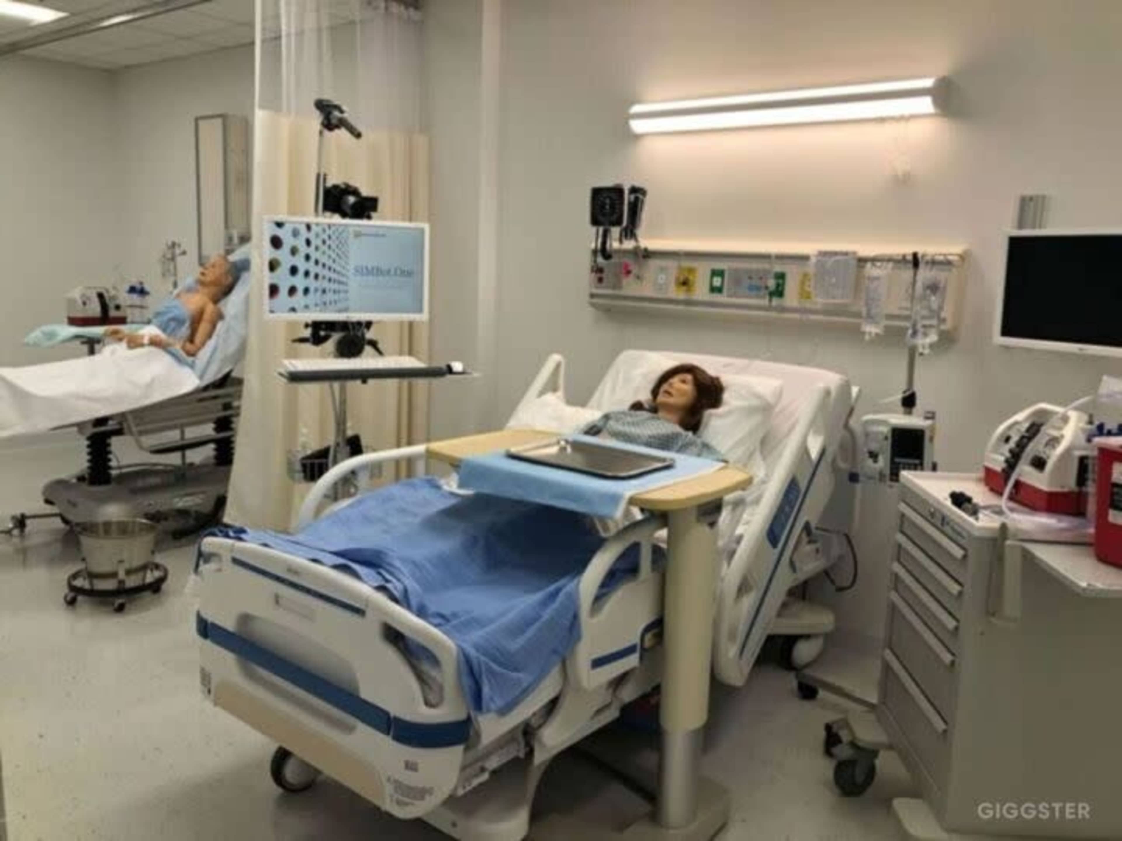 The image shows a hospital room equipped with two patient beds, one occupied by a medical training mannequin and the other by a human patient, along with medical equipment and a display screen.