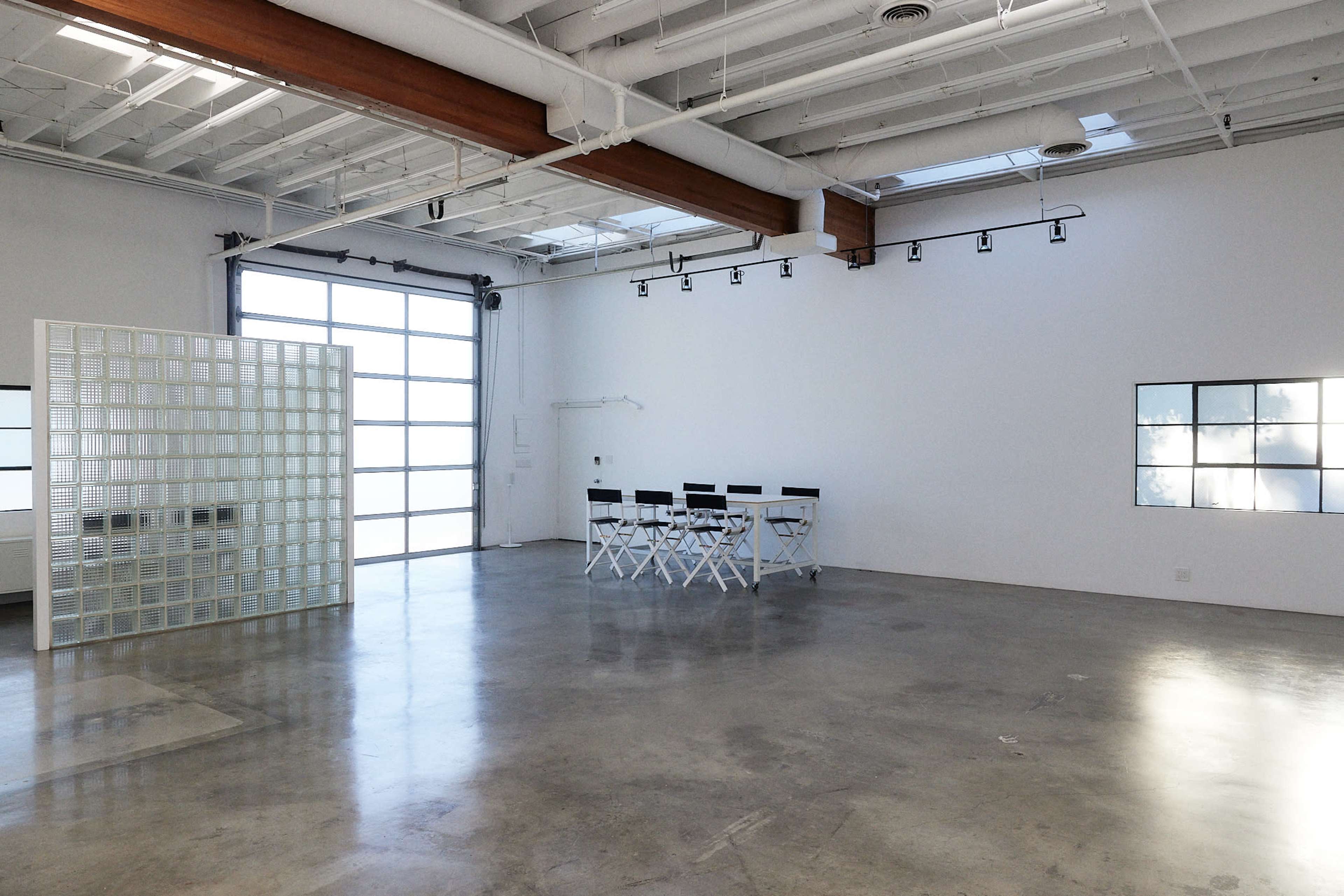 A spacious, modern room with concrete flooring, a glass block partition, and a row of white folding chairs against a wall of large windows.