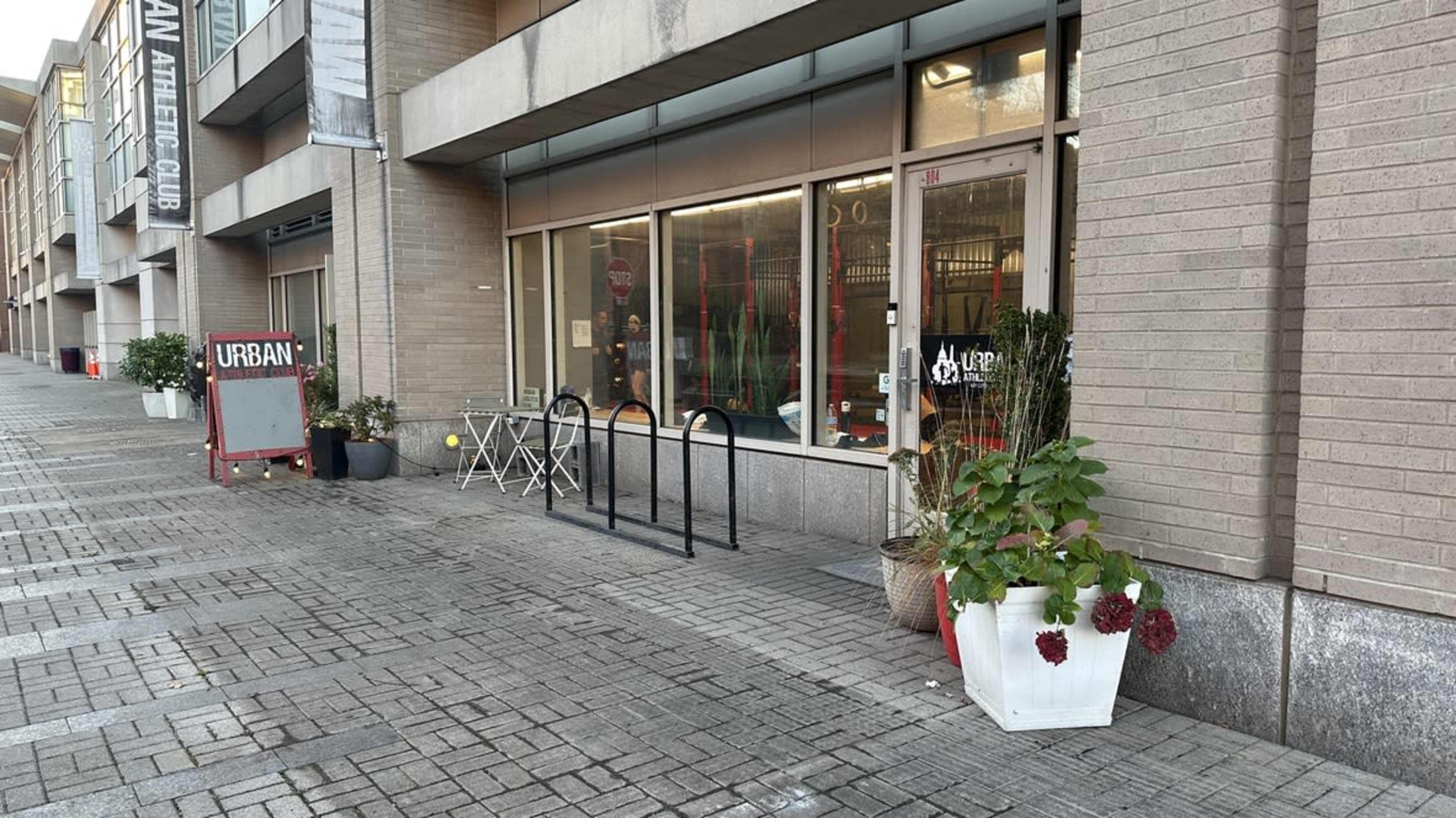The image shows a sidewalk area in front of a building with large windows, featuring outdoor seating with a small table and chairs, along with planters and a sign that reads "URBAN."