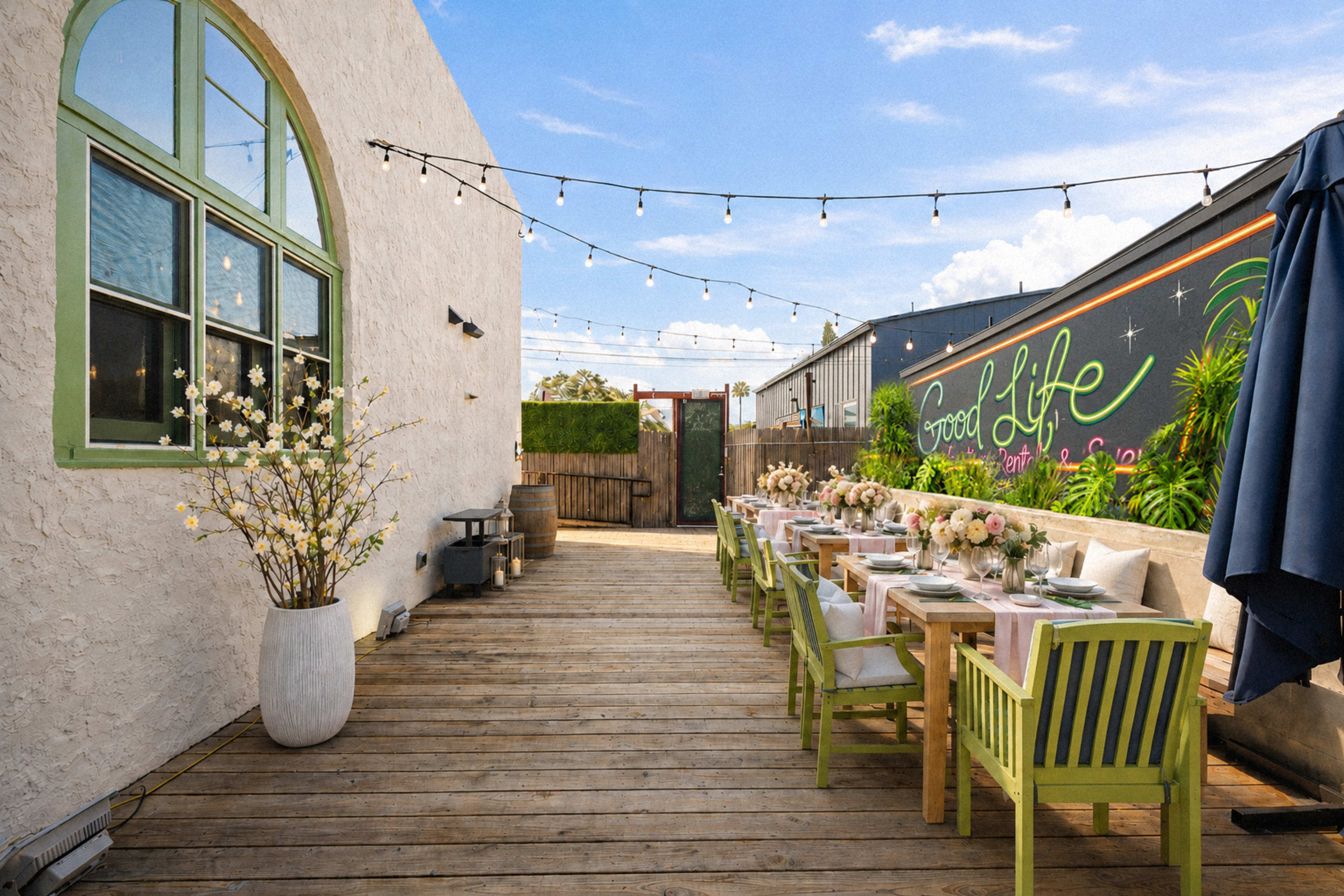 A long wooden dining table is set up outdoors on a patio, featuring flowers and greenery, with string lights overhead and a colorful mural in the background.