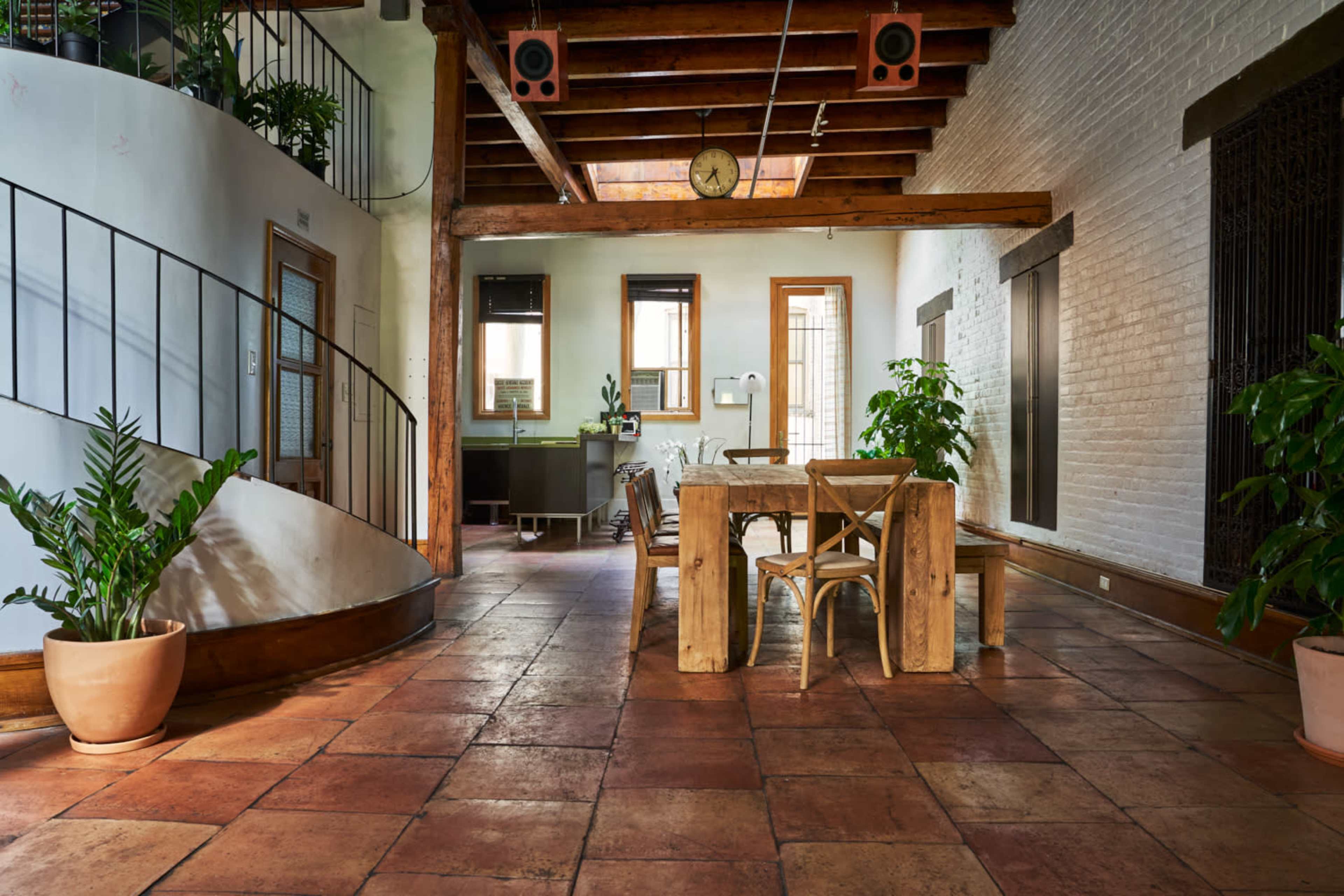 A spacious interior with exposed wooden beams, a large dining table, and potted plants, illuminated by natural light from several windows.