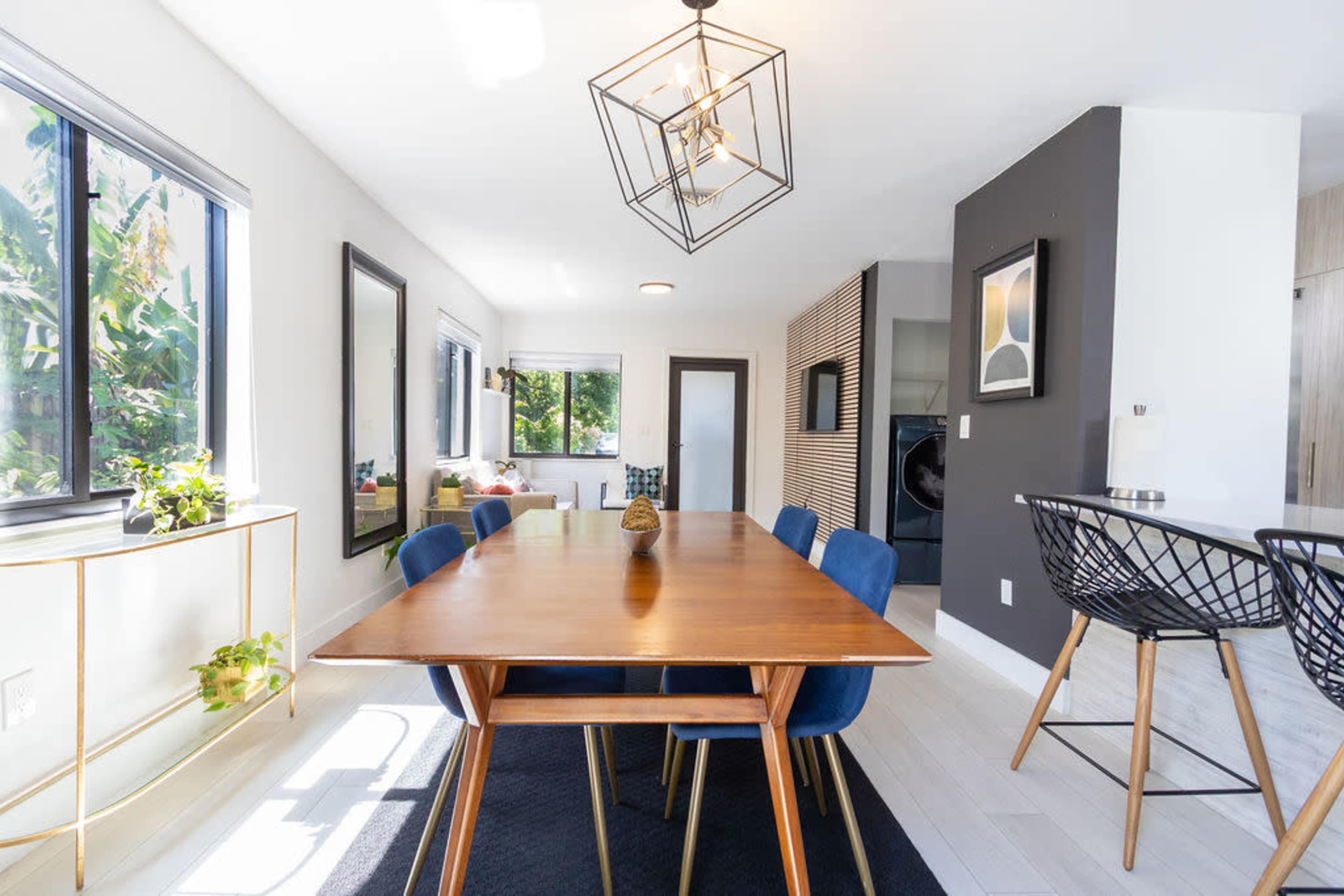 A modern dining area features a wooden table surrounded by blue chairs, with large windows providing natural light and plants enhancing the decor.