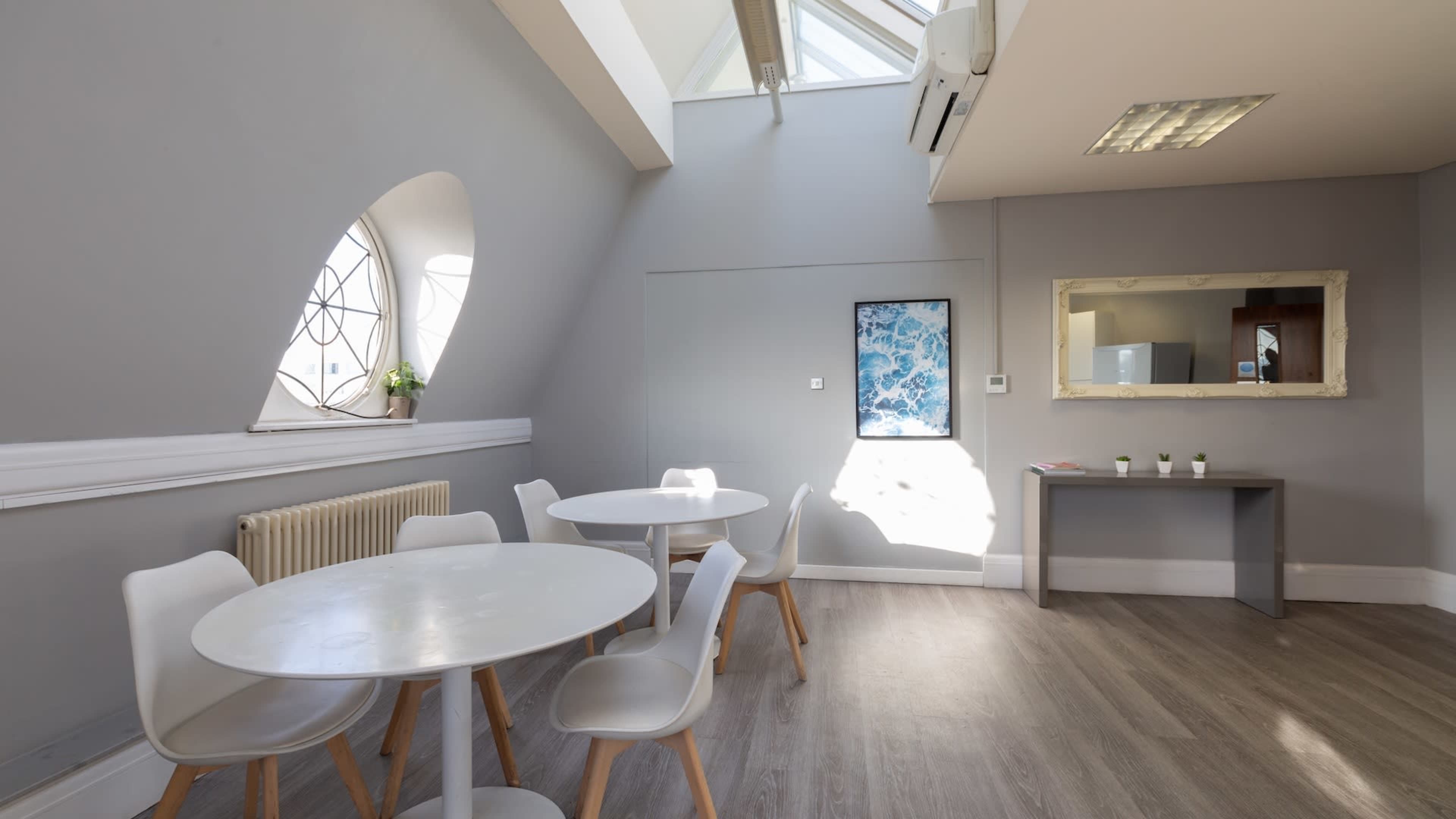 A modern, well-lit room features two round tables surrounded by white chairs, with a large window and a mirror on the walls.