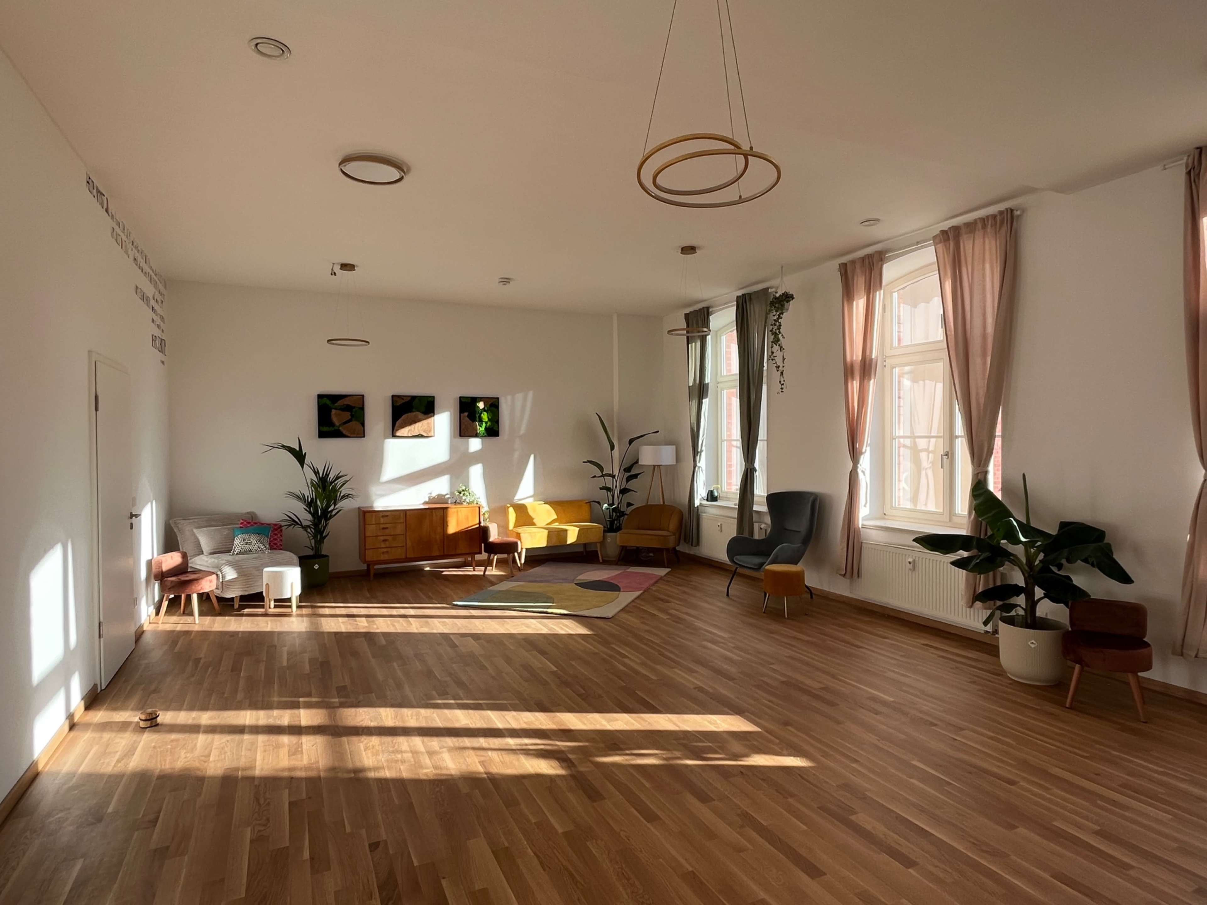 The image shows a spacious, sunlit room with wooden flooring, featuring various seating options and plants.