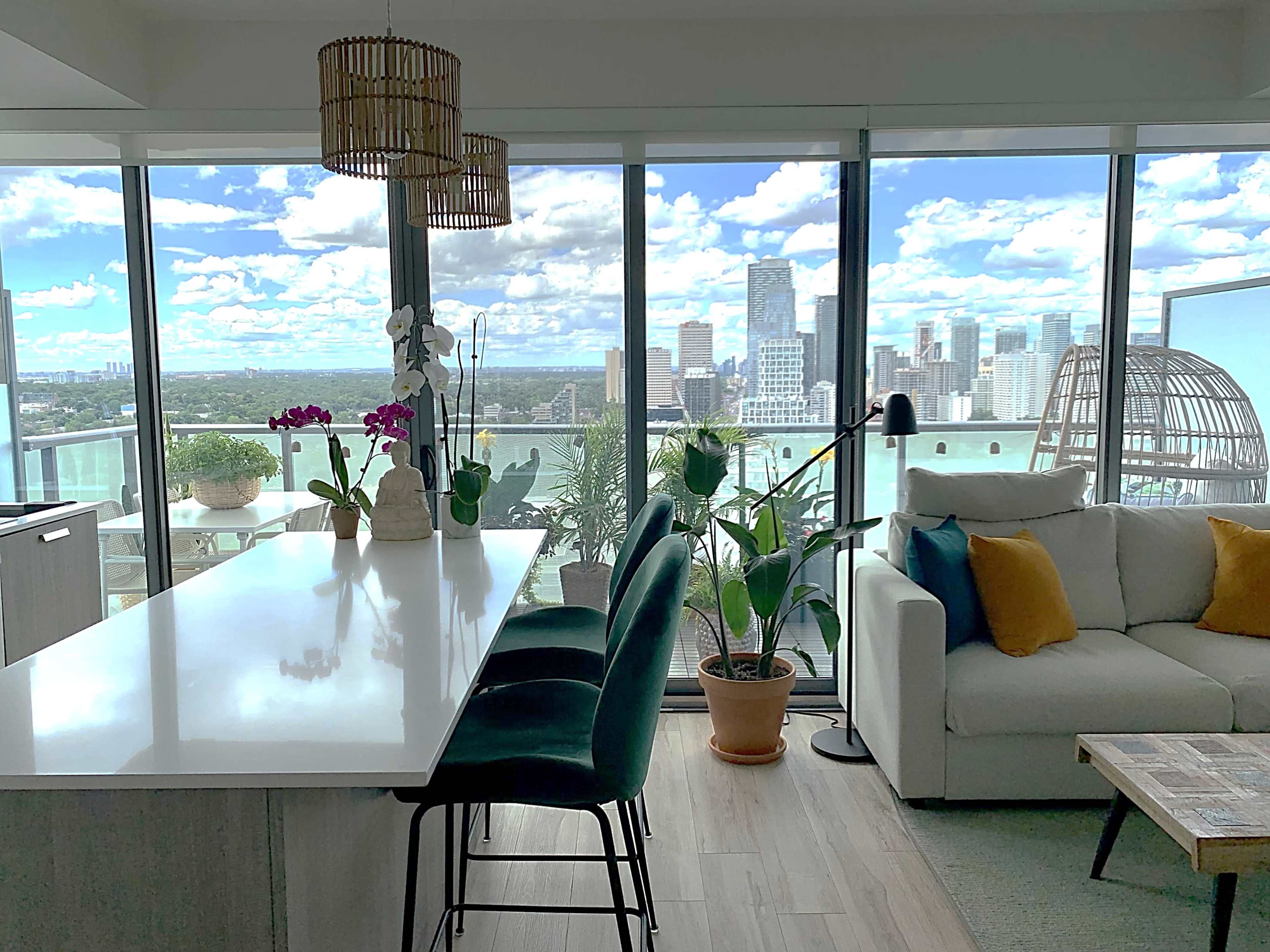 A modern living space features a kitchen island with bar stools, surrounded by large windows that showcase a city skyline and outdoor plants.