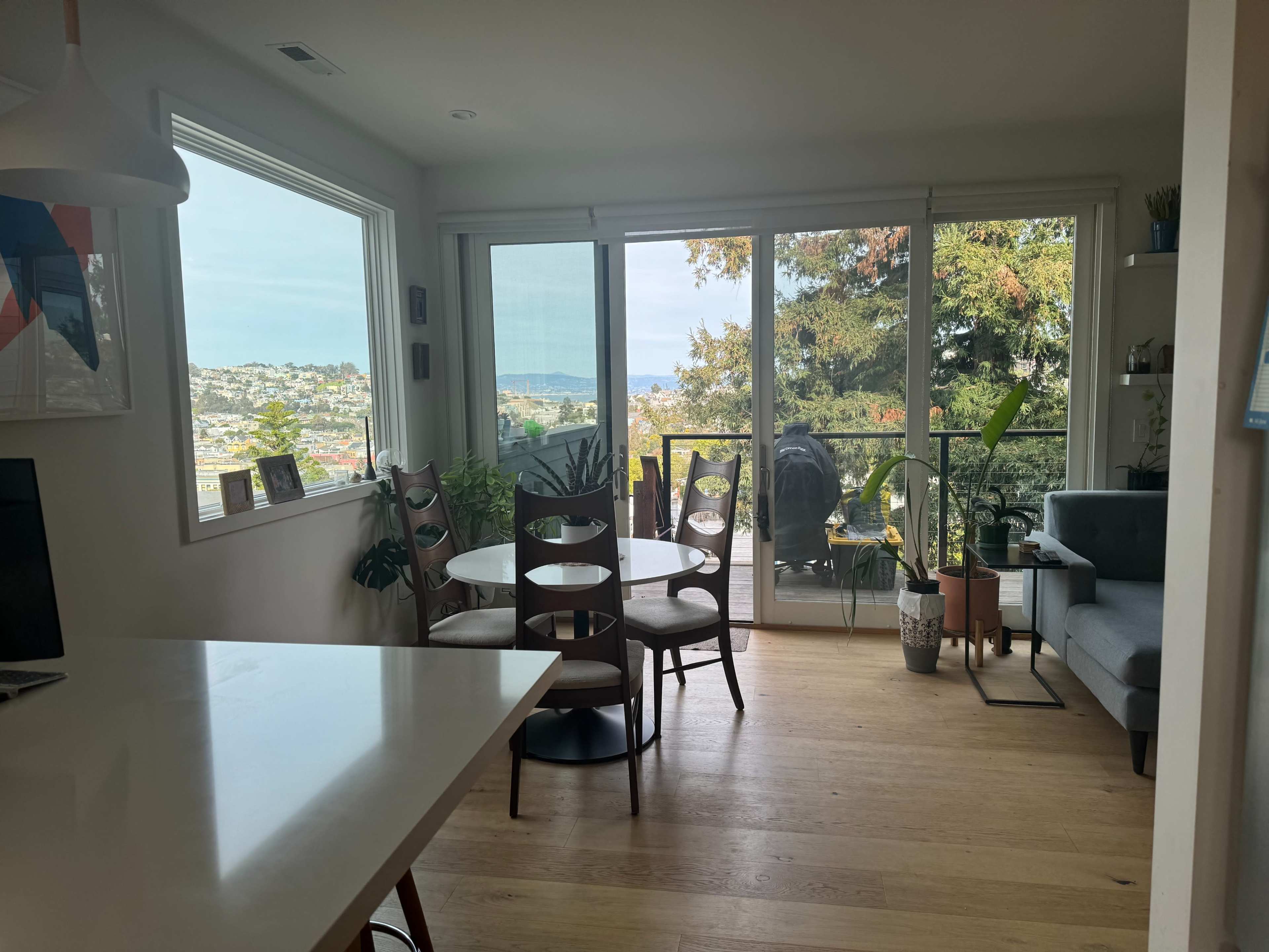 A bright living space featuring a dining area with wooden chairs, a modern couch, and large windows overlooking a scenic view with trees and water in the distance.